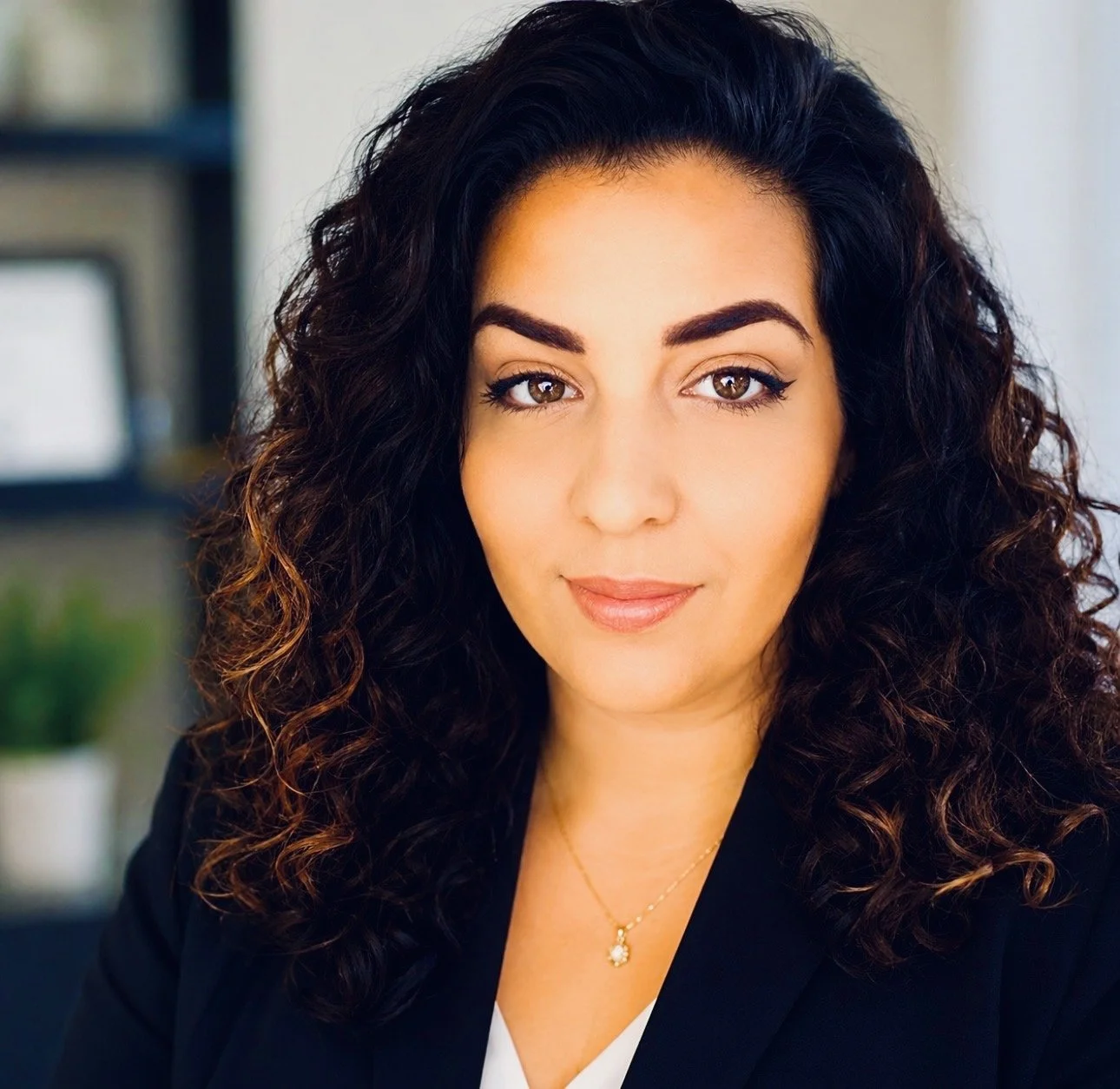 A woman with curly dark hair and fair skin, wearing a black blazer, white top, and a gold necklace, smiling softly in an indoor setting with a blurred background.