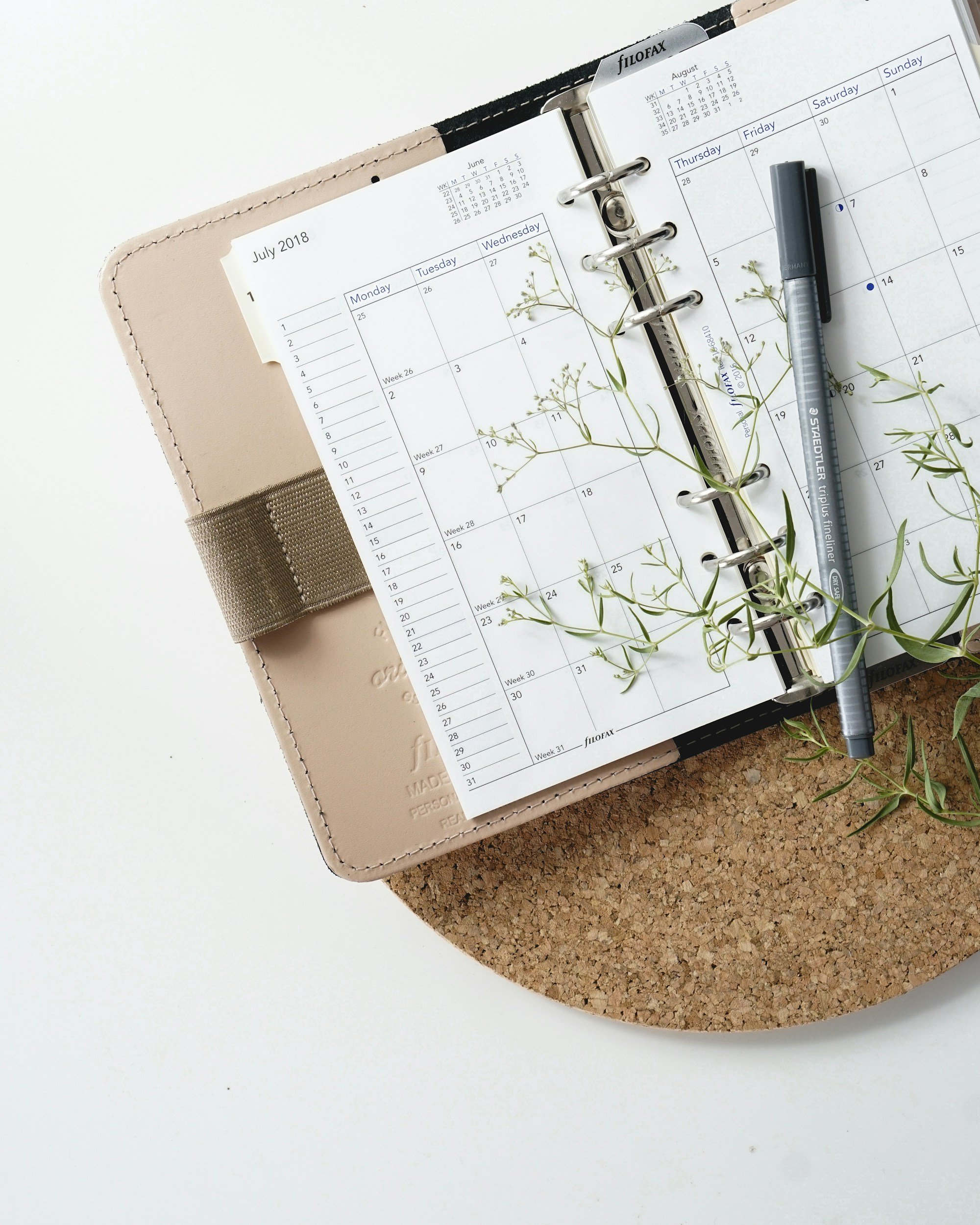 Open planner showing July 2018 and part of August 2018 calendar pages, with a gray marker and some sprigs of green plant on the pages, placed on a two-tone surface with white and cork textures.