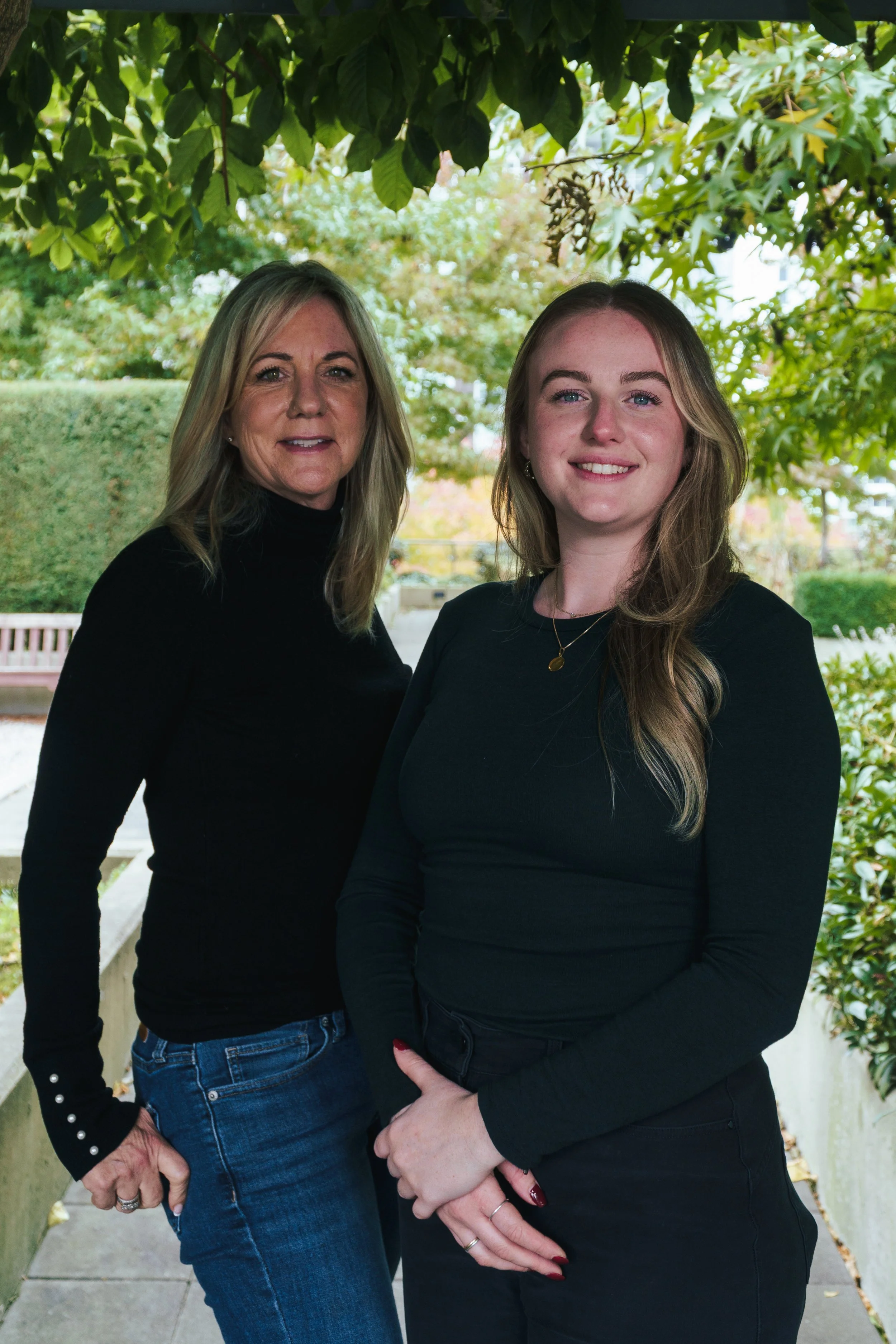 Two women founders of Kindred Memorials (Leah and Courtenay), standing close together outdoors under tree branches with green leaves, smiling at the camera.
