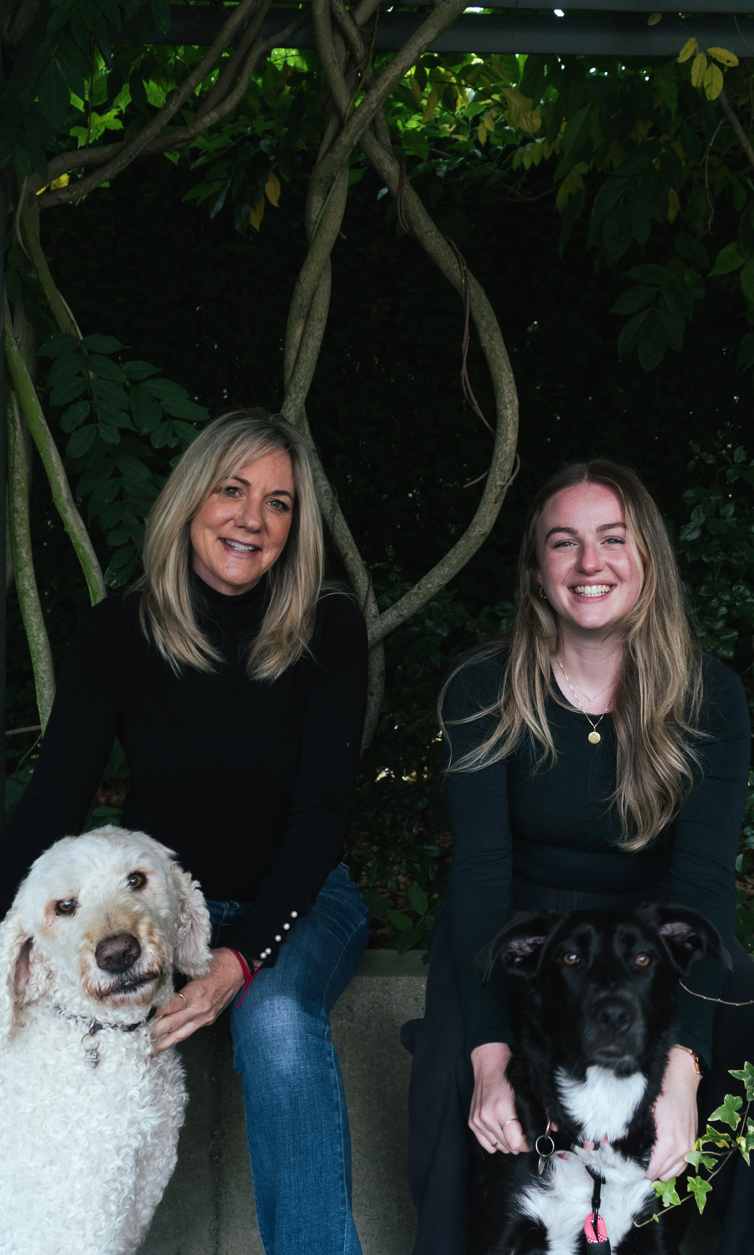 Two women sitting outdoors with two dogs, a white dog with curly fur and a black and white dog, in front of green leafy bushes.