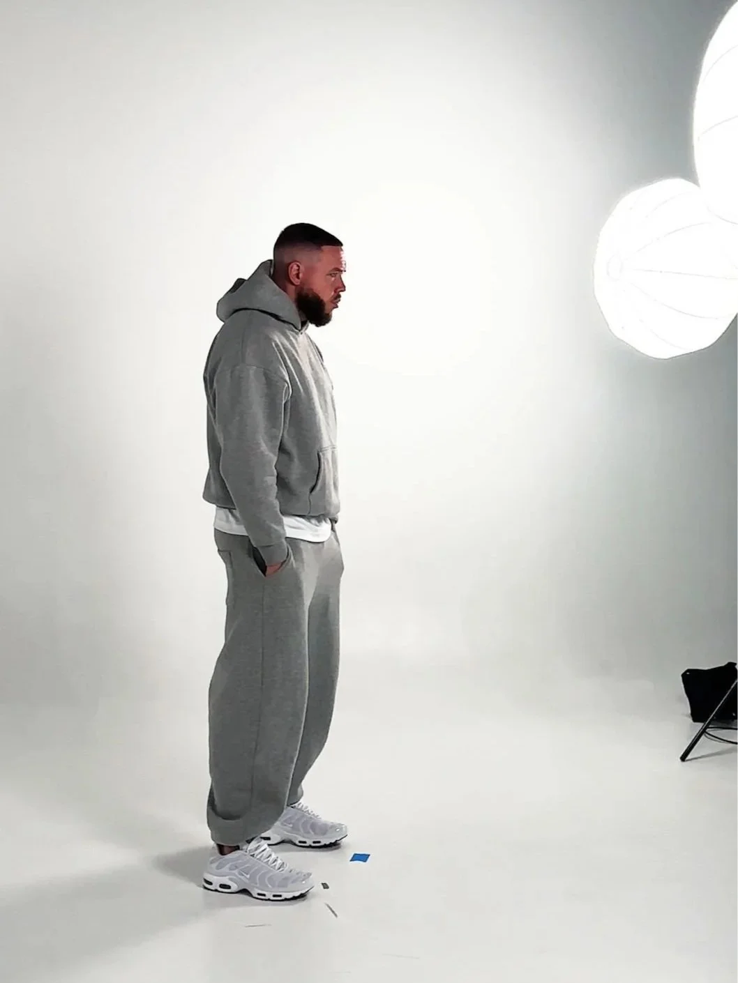 Man standing in a white photography studio, wearing gray hoodie, gray sweatpants, and white sneakers, with studio lights overhead.