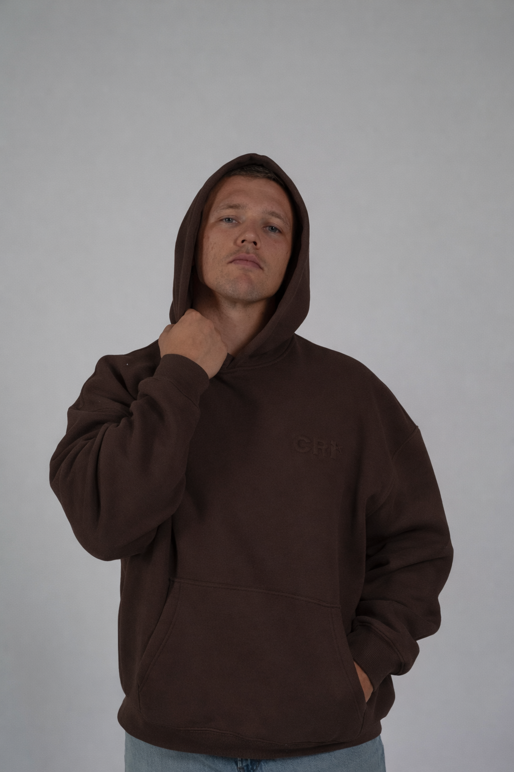 A young man wearing a brown hoodie with the hood up, standing against a plain, light-colored background. He has short hair and a serious expression, with his left hand in his pocket and his right hand touching his neck.
