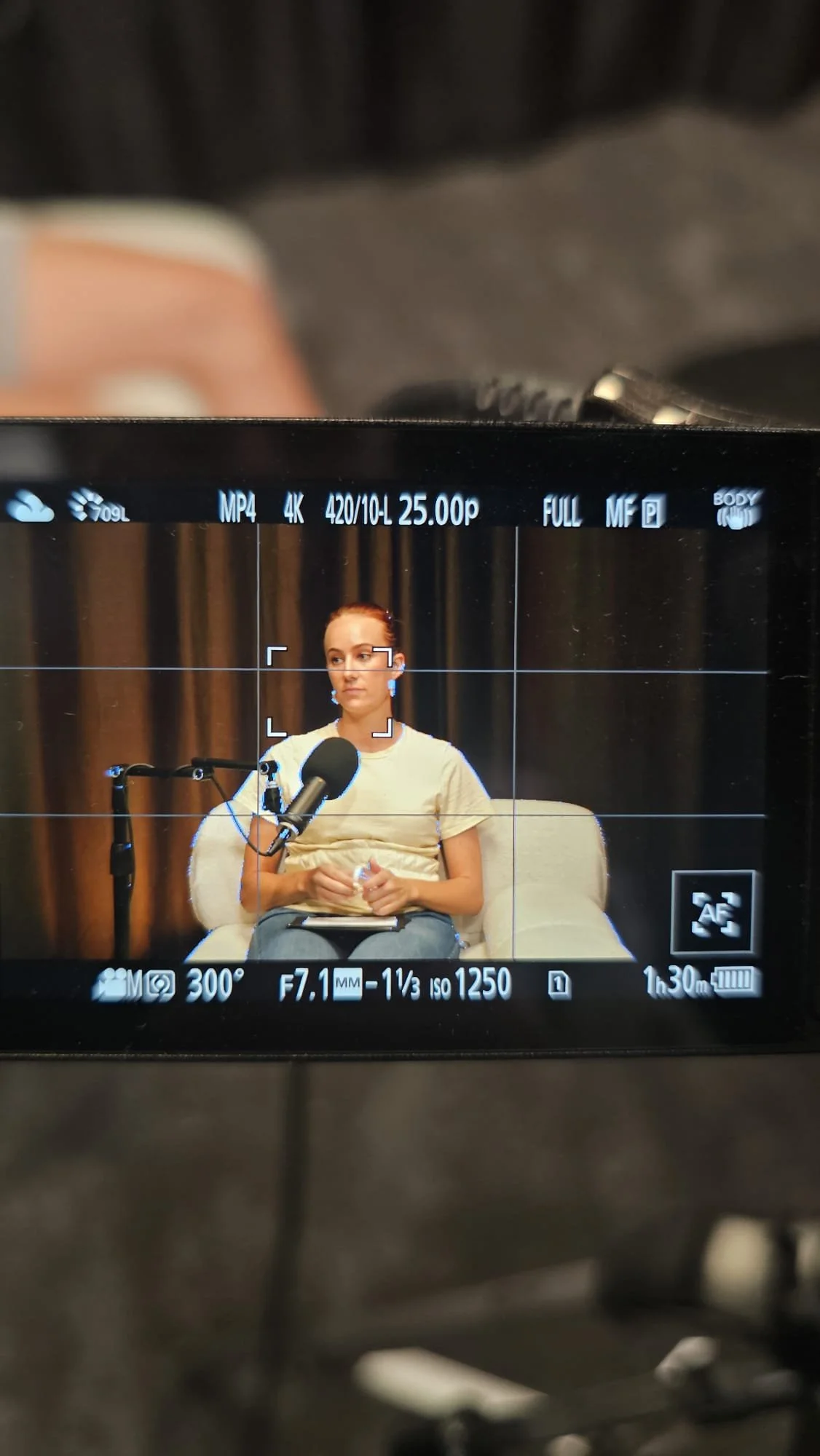A woman with red hair sitting on a beige couch, holding a water bottle, during a recorded or live video interview with a microphone in front of her, with a dark curtain background.
