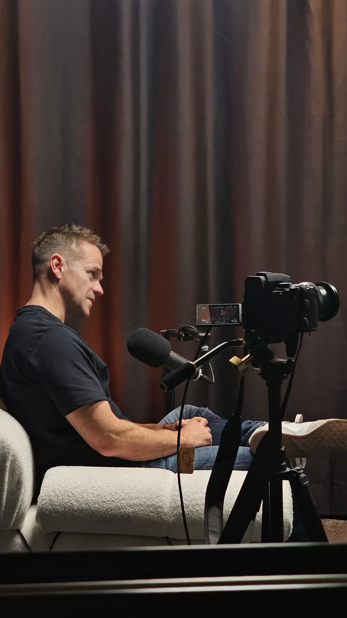 A man sitting on a white couch in front of a professional camera setup with a microphone, in a room with dark curtains.
