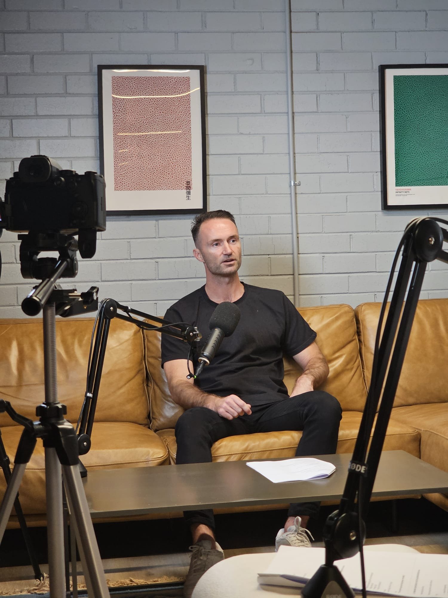 A man sitting on a yellow leather couch in a room with white brick walls, surrounded by filming equipment, with framed art on the wall behind him.