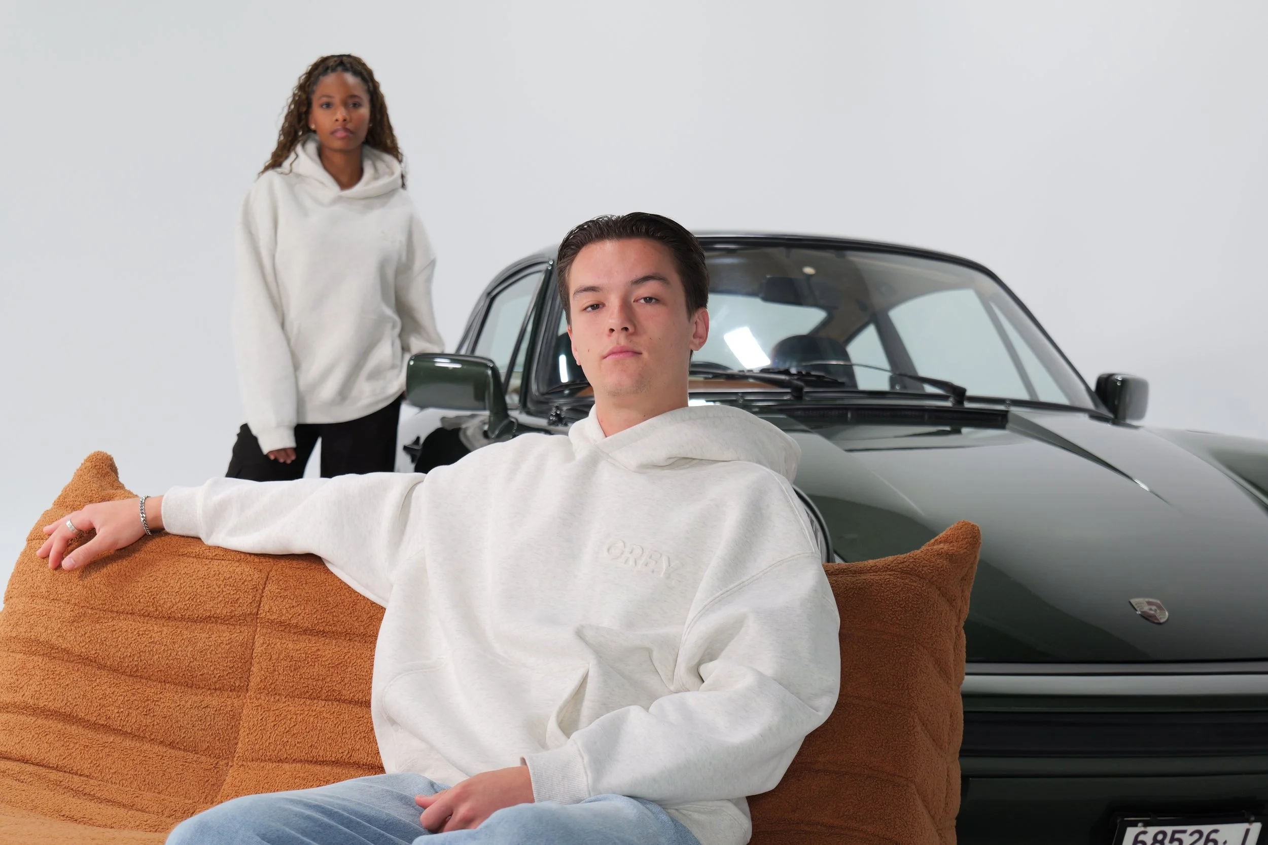 A young man sitting on a brown sofa with a relaxed pose, wearing a white hoodie and blue jeans, in front of a black vintage car. Behind him is a young woman in a white hoodie, standing near the car, with a neutral expression. The setting has a plain white wall background.