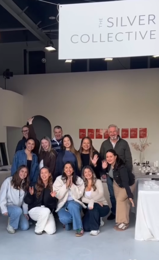 Group of people smiling and waving at an event called The Silver Collective, with tables and decorations in the background.