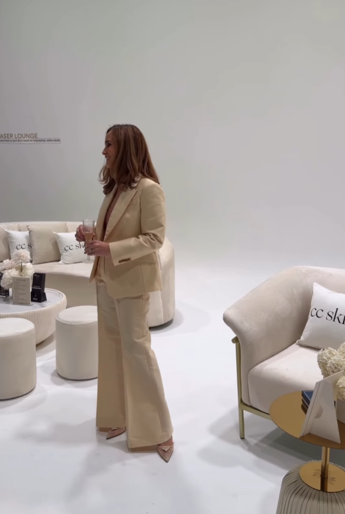 Woman in beige suit standing in a modern, minimalist lounge area holding a glass of champagne.