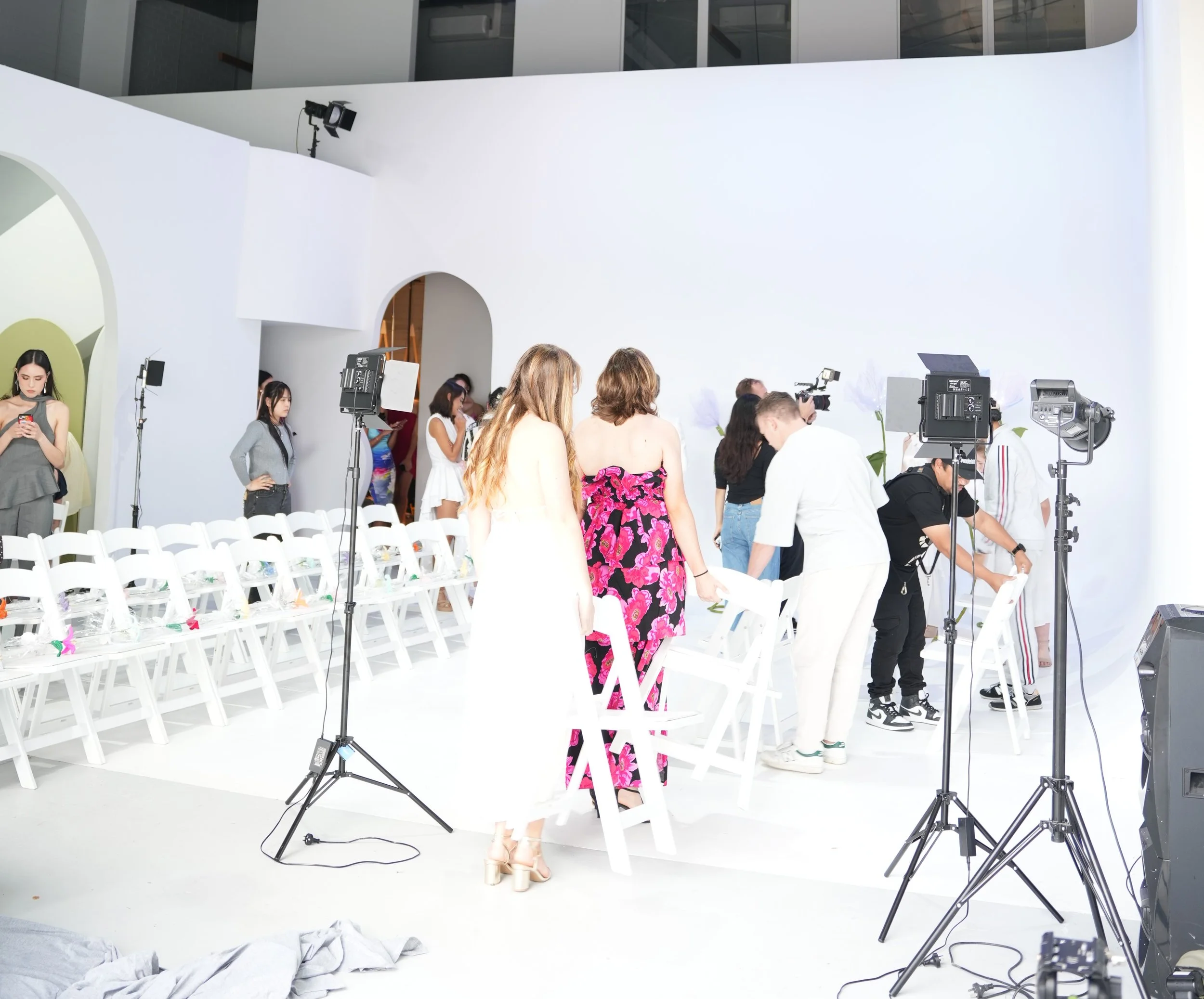 People preparing for a photo shoot in a white studio with professional lighting equipment. Women are dressed in stylish outfits, and chairs are arranged in front of the backdrop.