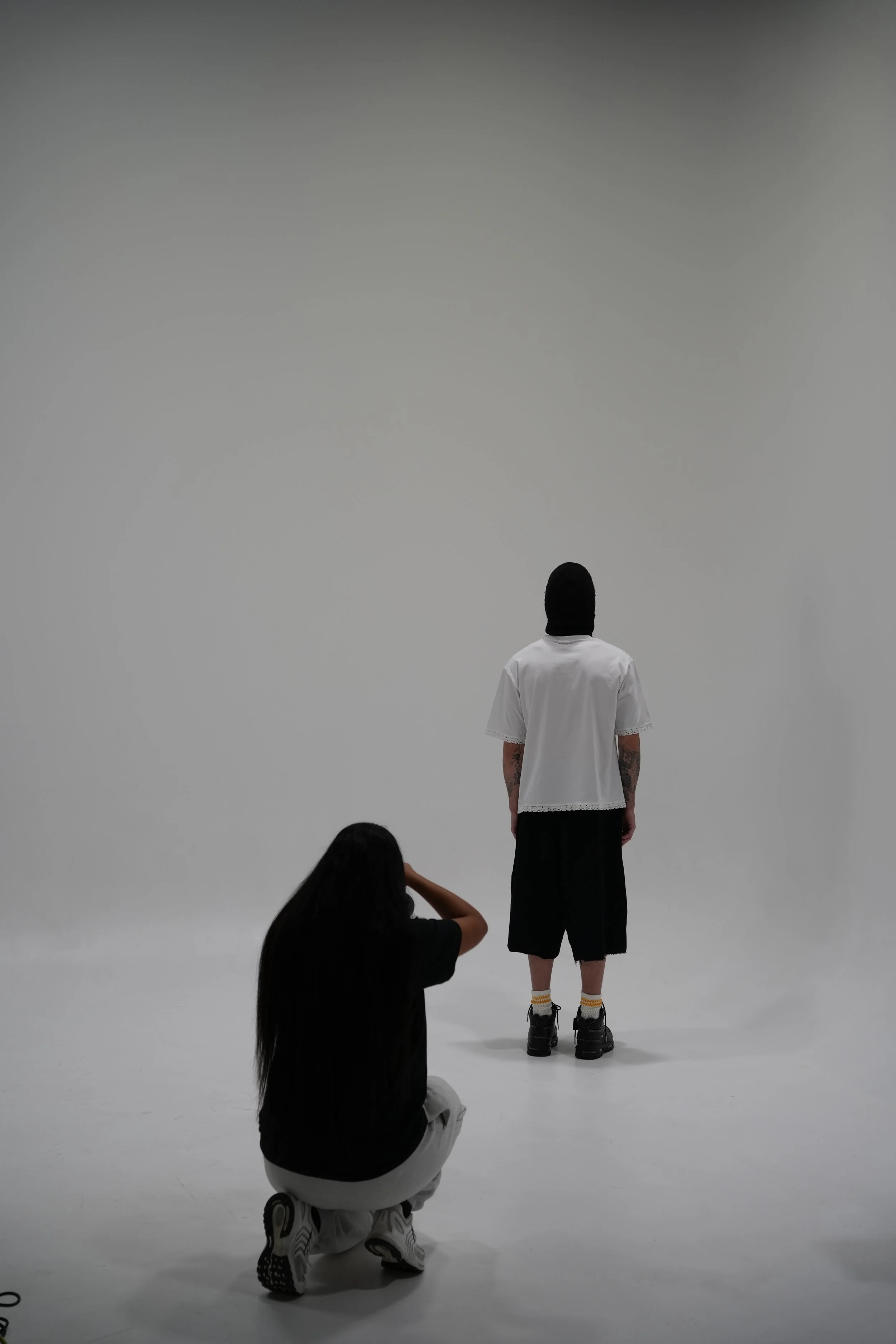 A person with tattoos on arms and wearing a white shirt and black shorts standing on a white studio floor, facing a plain light gray wall. Two women are in foreground; one is crouching, the other is sitting and taking a photo, both observing the person.