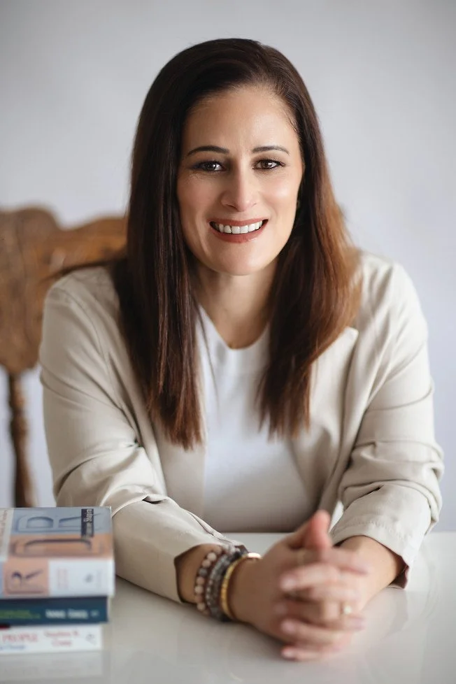A woman with long brown hair smiling and sitting at a table, wearing a cream-colored blazer, with books stacked on the table in front of her.