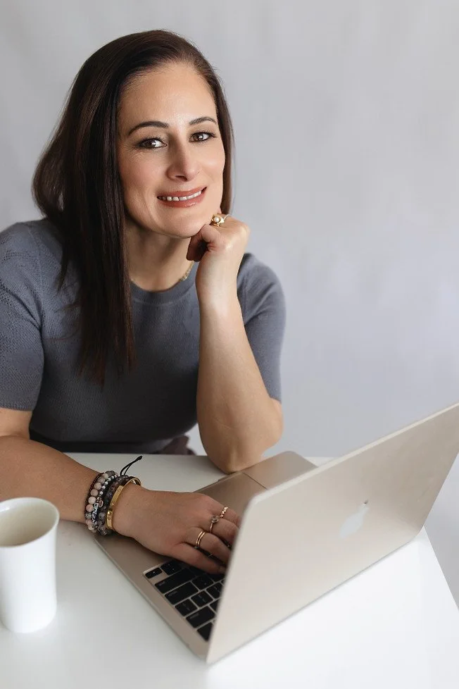 A woman with shoulder-length dark brown hair sitting at a white table with a silver MacBook laptop, a white mug, and some jewelry, smiling and resting her chin on her hand.