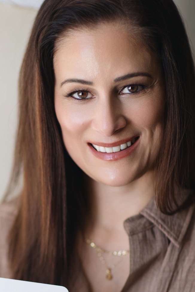 Close-up of smiling woman with brown hair and brown eyes wearing a brown shirt and gold necklace.