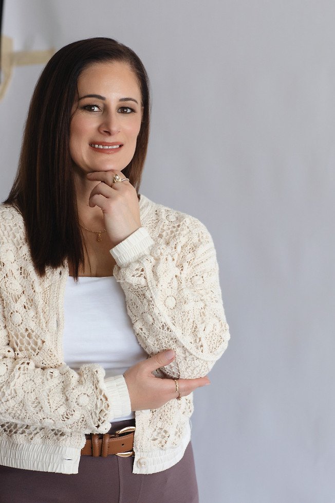 A woman with shoulder-length brown hair, smiling, posing with her chin resting on her right hand and her left hand resting on her stomach. She is wearing a white top, a cream-colored knitted cardigan, and brown pants, standing against a plain light gray wall.