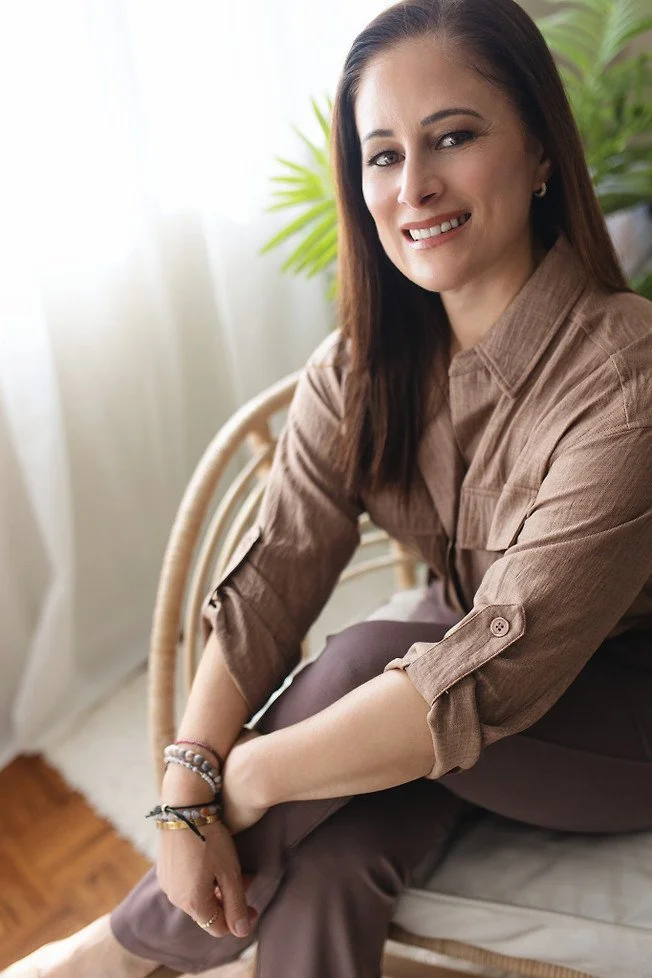 A woman with brown hair sitting on a chair, smiling at the camera, with a green plant and bright window in the background.