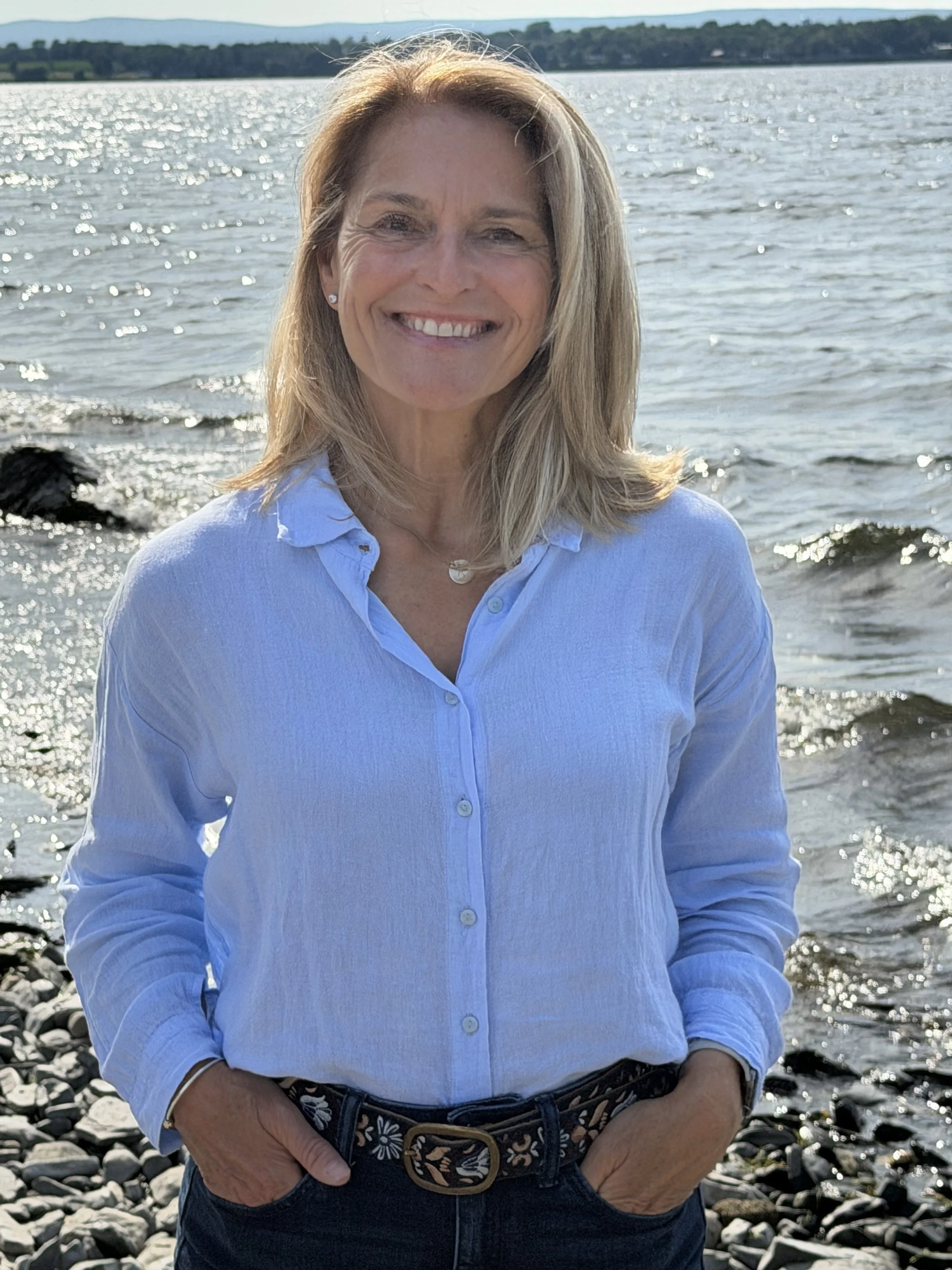 A woman standing on a rocky shoreline near a body of water with a distant tree-lined shore in the background, smiling at the camera.