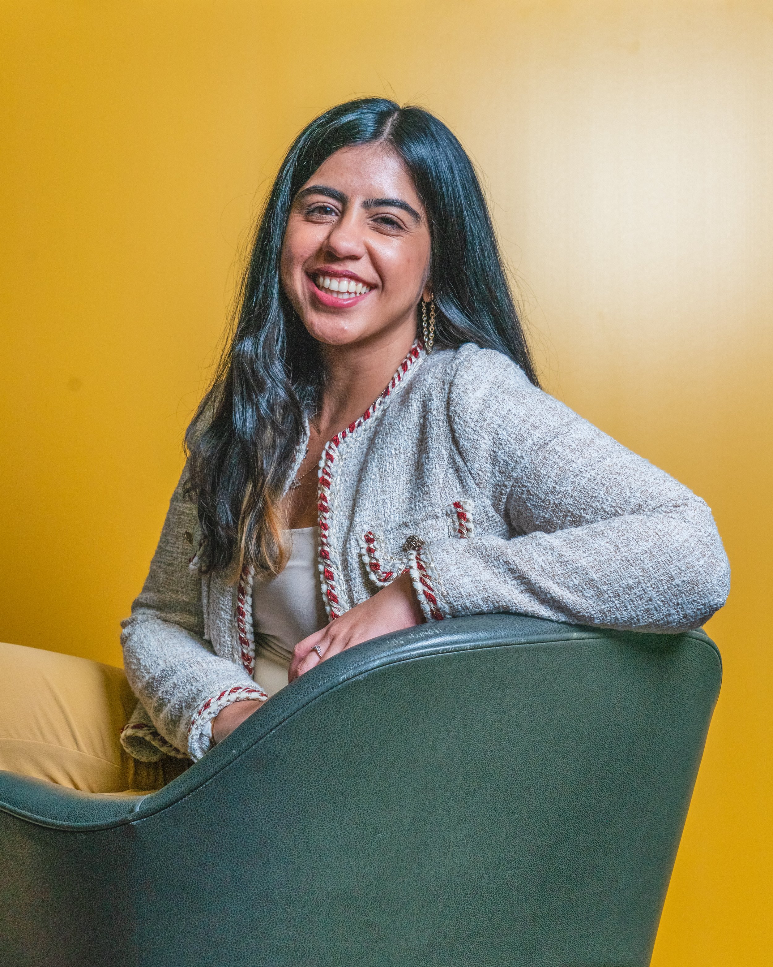 Sanam Panjwani, a South Asian yoga practitioner and wellness coach in Toronto, smiling warmly in a relaxed seating pose against a vibrant yellow background, representing somatic movement and nervous system regulation.