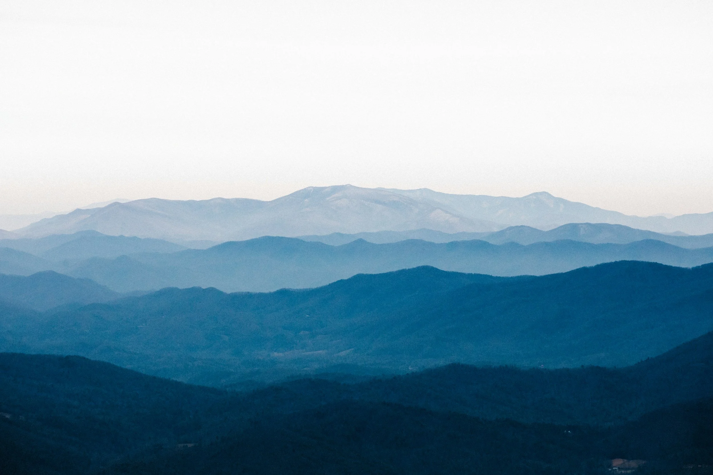 A landscape of multiple mountain ranges with a soft blue hue, fading into the distance under a pale sky.