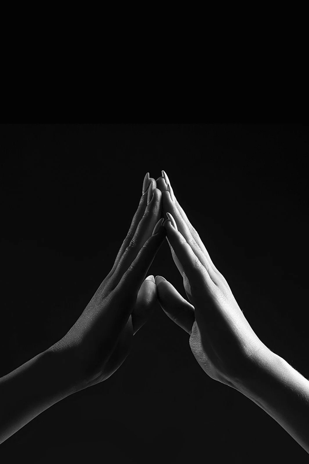 Black and white photo of two hands touching in prayer or a gesture of unity, with fingers pointed upward and palms pressed together, against a dark background.