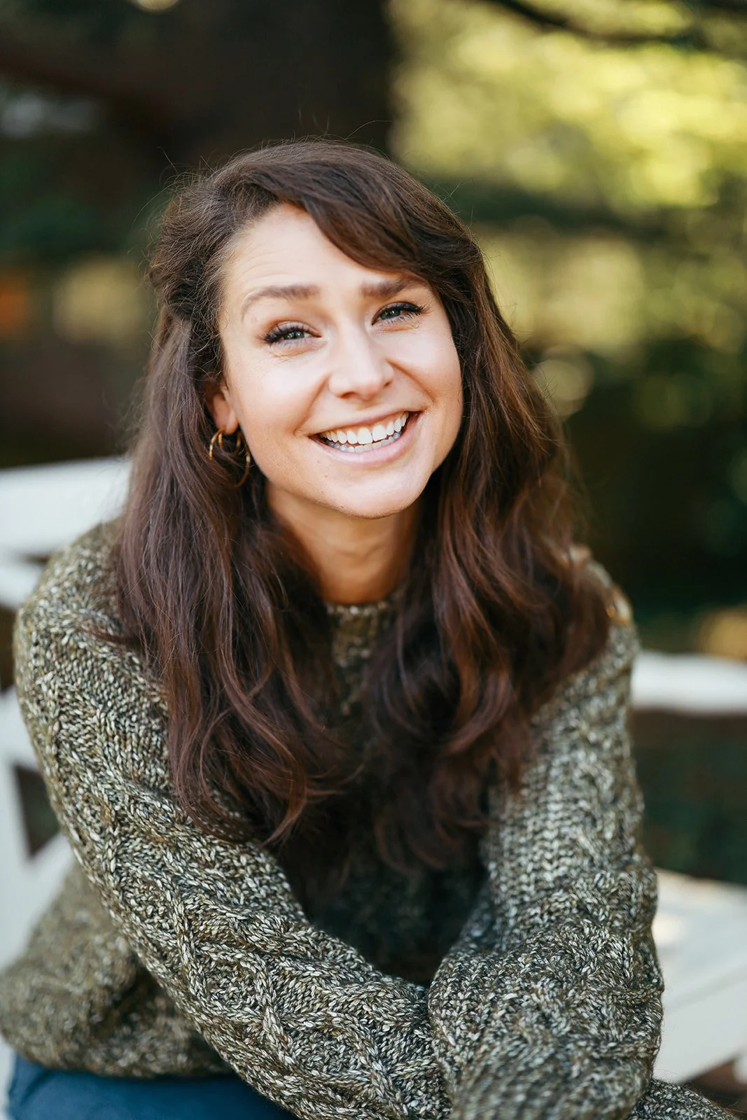 Headshot of a smiling woman with long brown wavy hair, wearing a patterned sweater, outdoors with blurred greenery in the background.