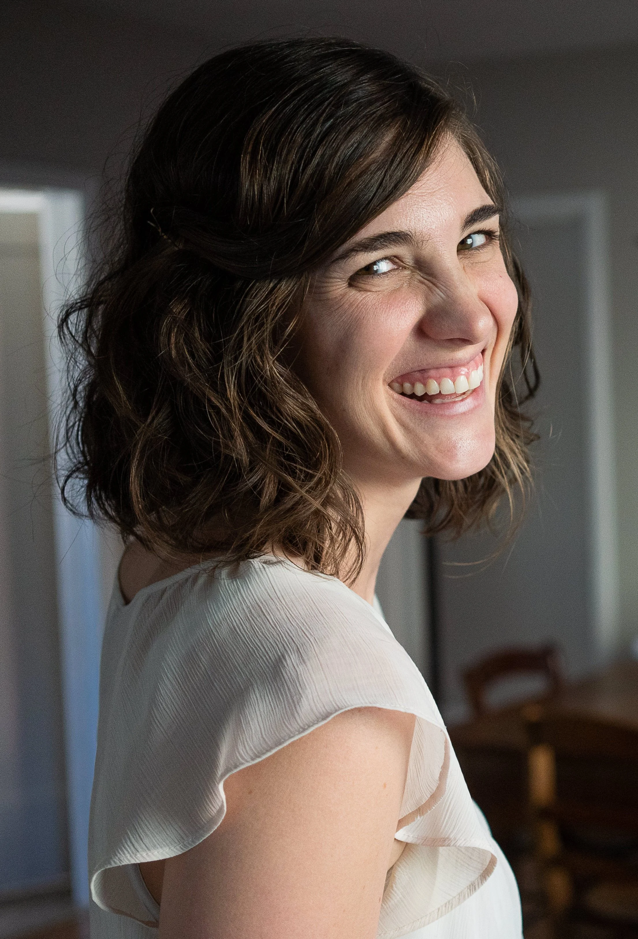 Smiling woman with wavy brown hair wearing a white ruffled sleeve top, indoors.
