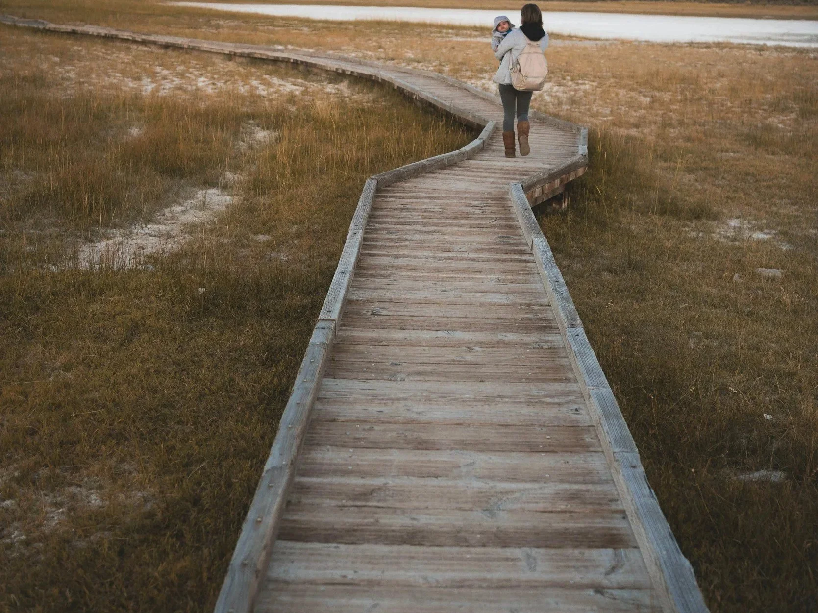 A woman walking along a winding wooden boardwalk in a marshland, carrying a child in her arms.