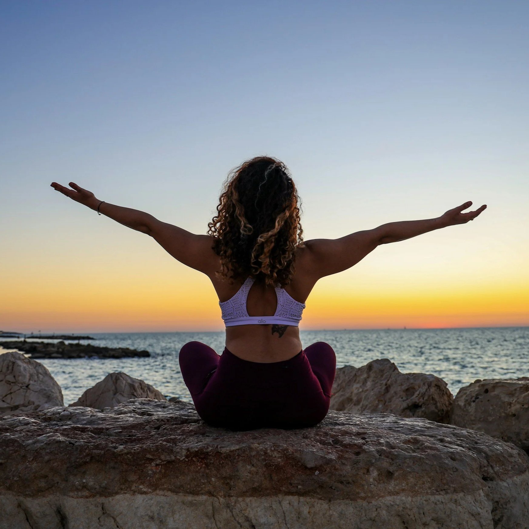 A woman practicing yoga or meditation on a rocky shoreline at sunset or sunrise, with her arms outstretched and overlooking the ocean.