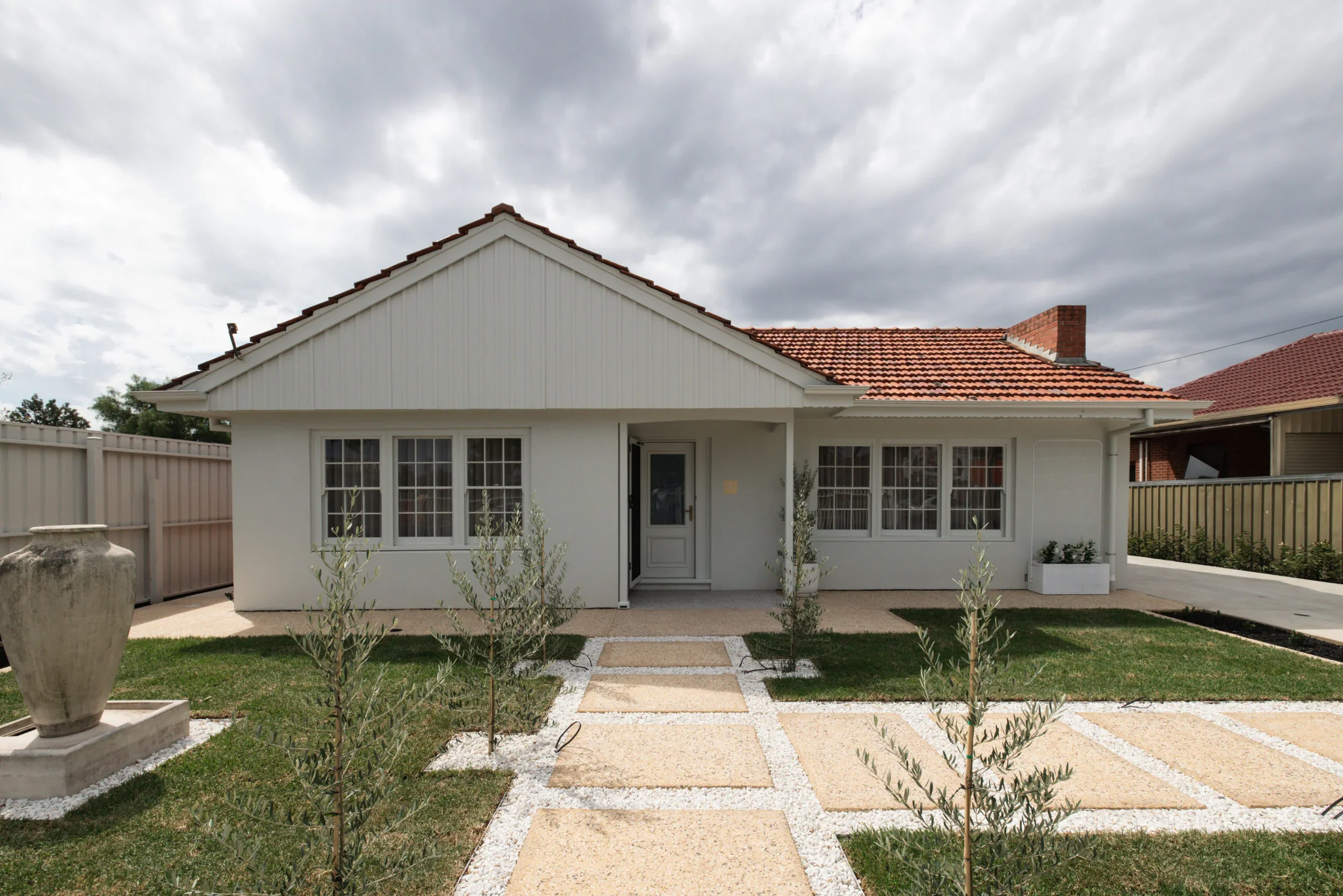 Front view of a residential house with a red-tiled roof, white exterior walls, large windows, and a small front yard with a pathway, plants, and a decorative planter. Overcast sky with dark clouds.