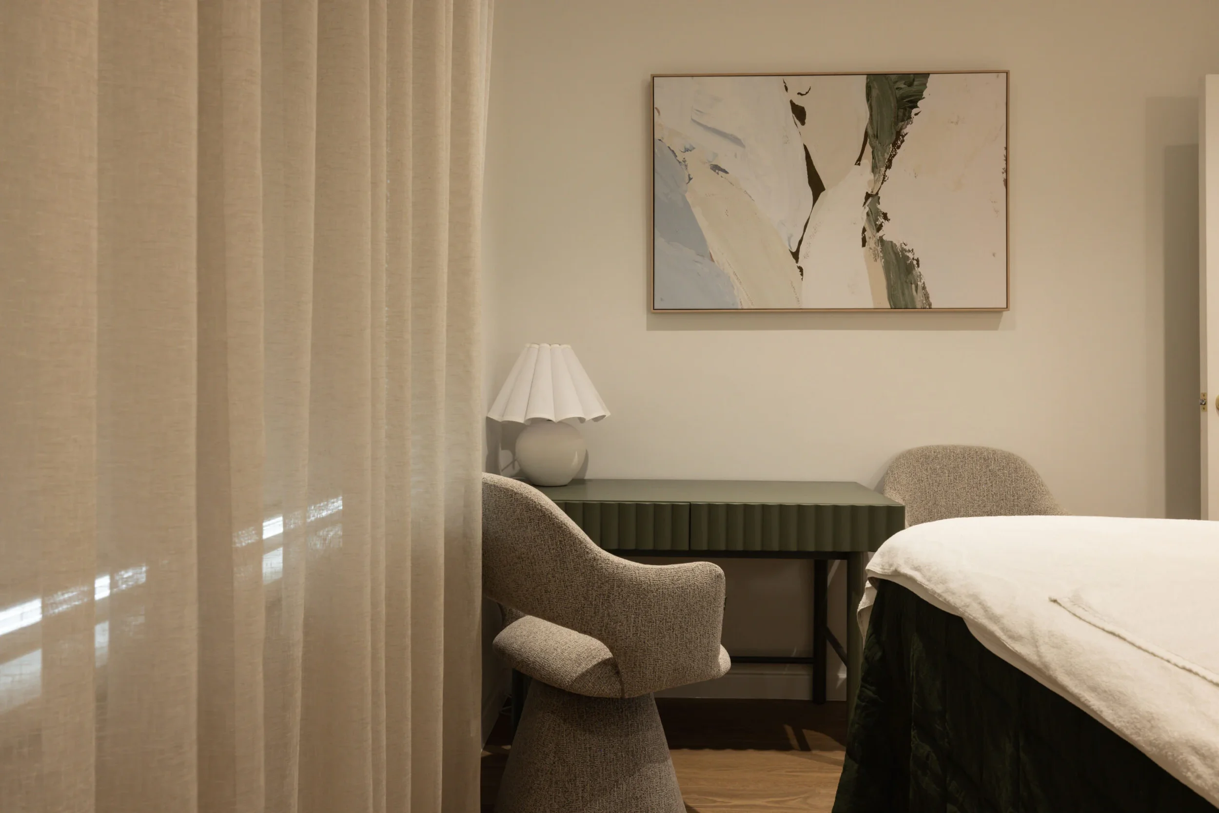 A cozy bedroom corner with beige curtains, a white table lamp, a decorative table, and an abstract landscape painting on the wall.