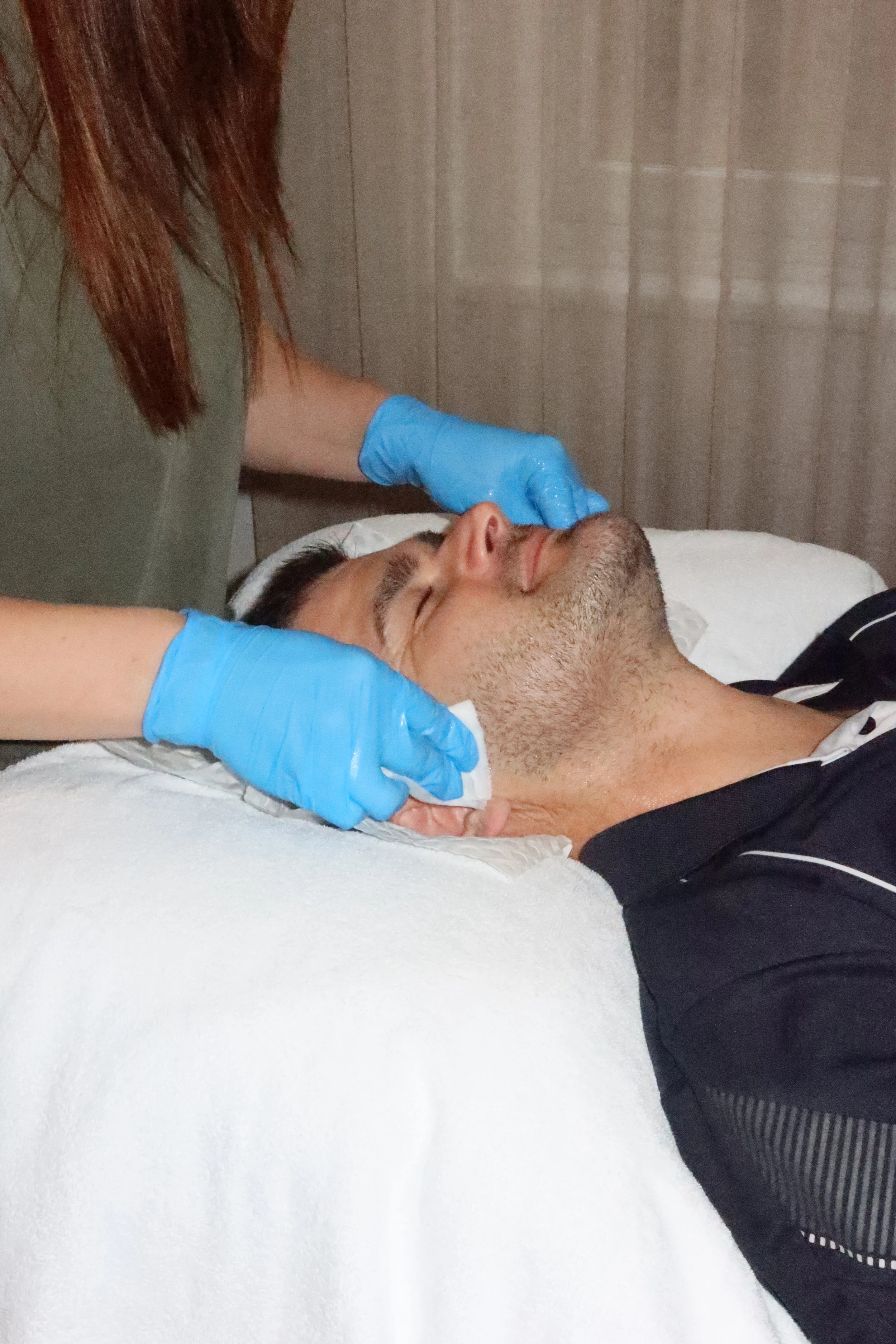 A person lying on a bed with eyes closed, receiving care from a second person wearing blue gloves, who is gently touching the face of the person on the bed.