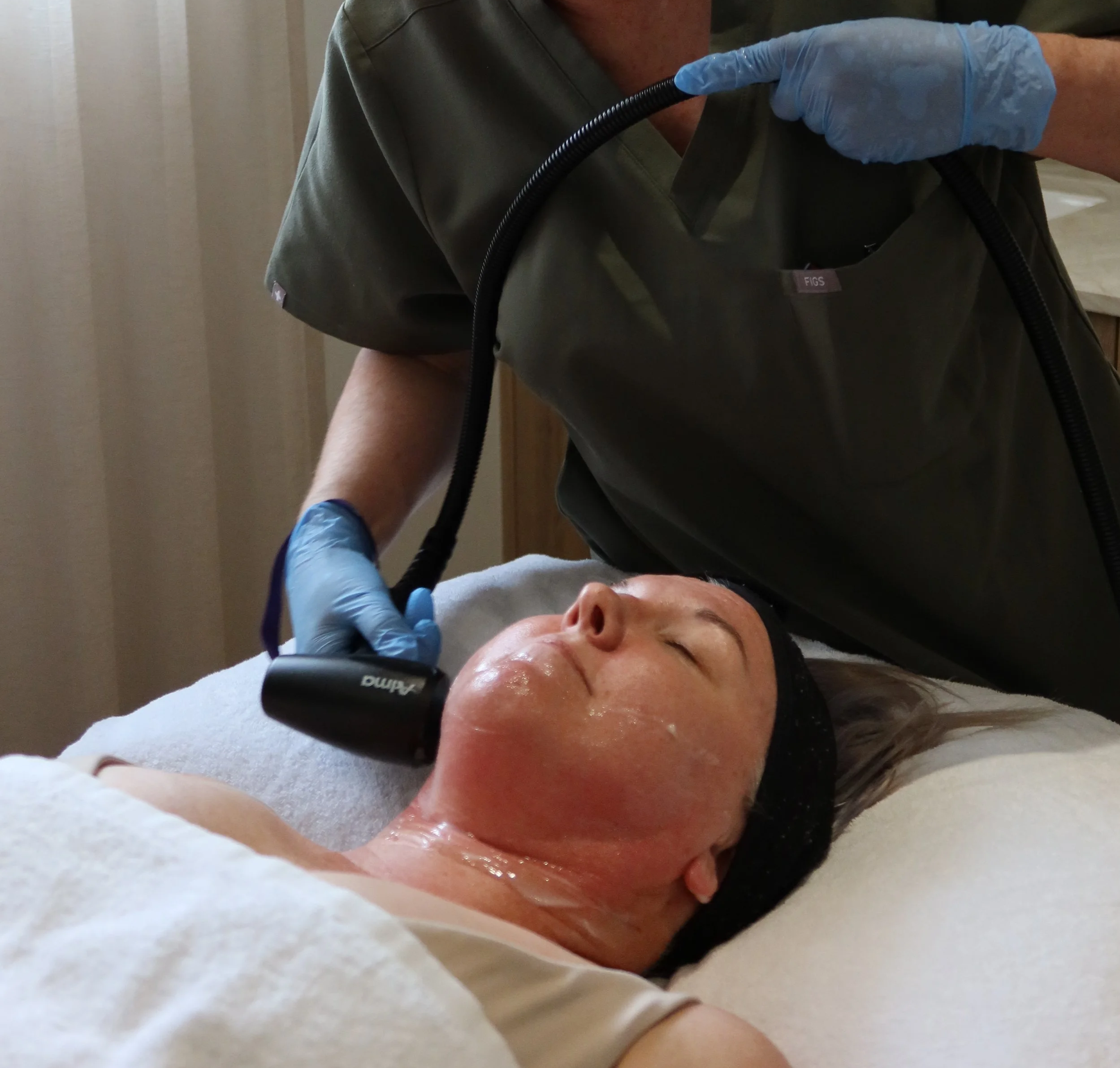 Close-up of a woman receiving a facial treatment, lying on a white towel with her eyes closed, a healthcare worker wearing blue gloves and a green medical uniform standing beside her, using a handheld device.