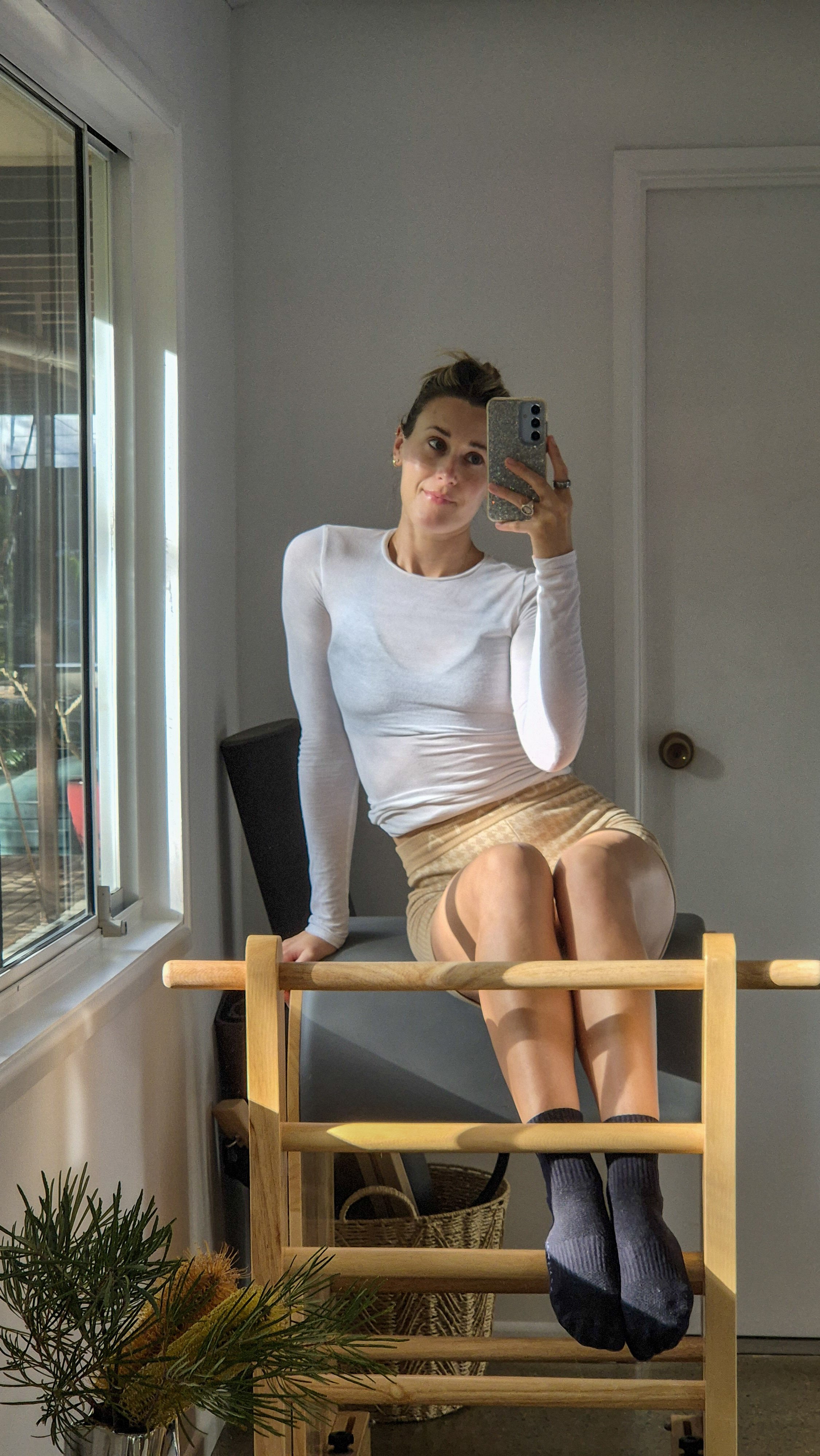 A woman sitting on a chair taking a mirror selfie with her smartphone in a room with a window and a door.