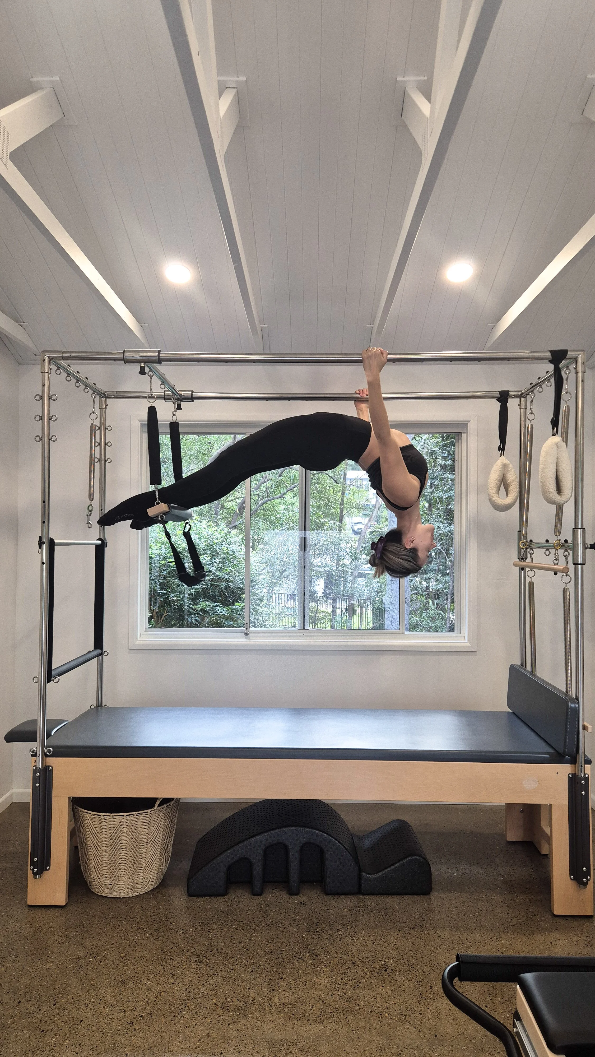 A woman in black workout clothes performing an inverted form on a wall-mounted Pilates Cadillac trapeze table, inside a gym with a window showing trees outside.