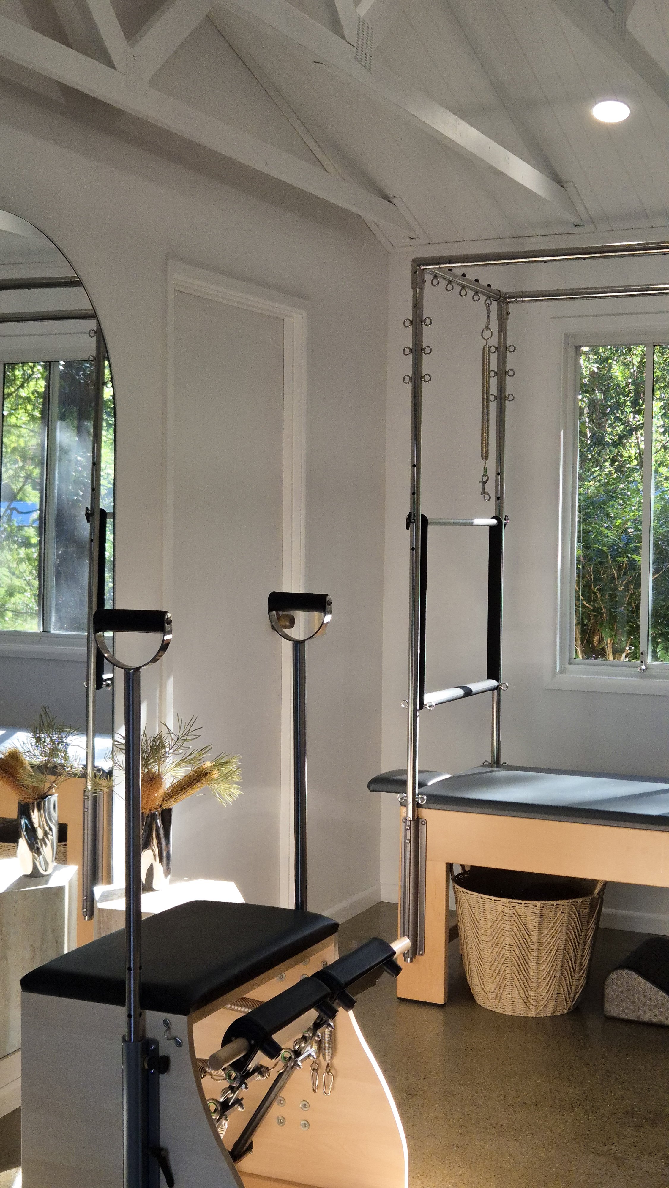 A room with Pilates equipment, including a reformer and a Cadillac, next to windows with greenery outside, sunlight streaming in.