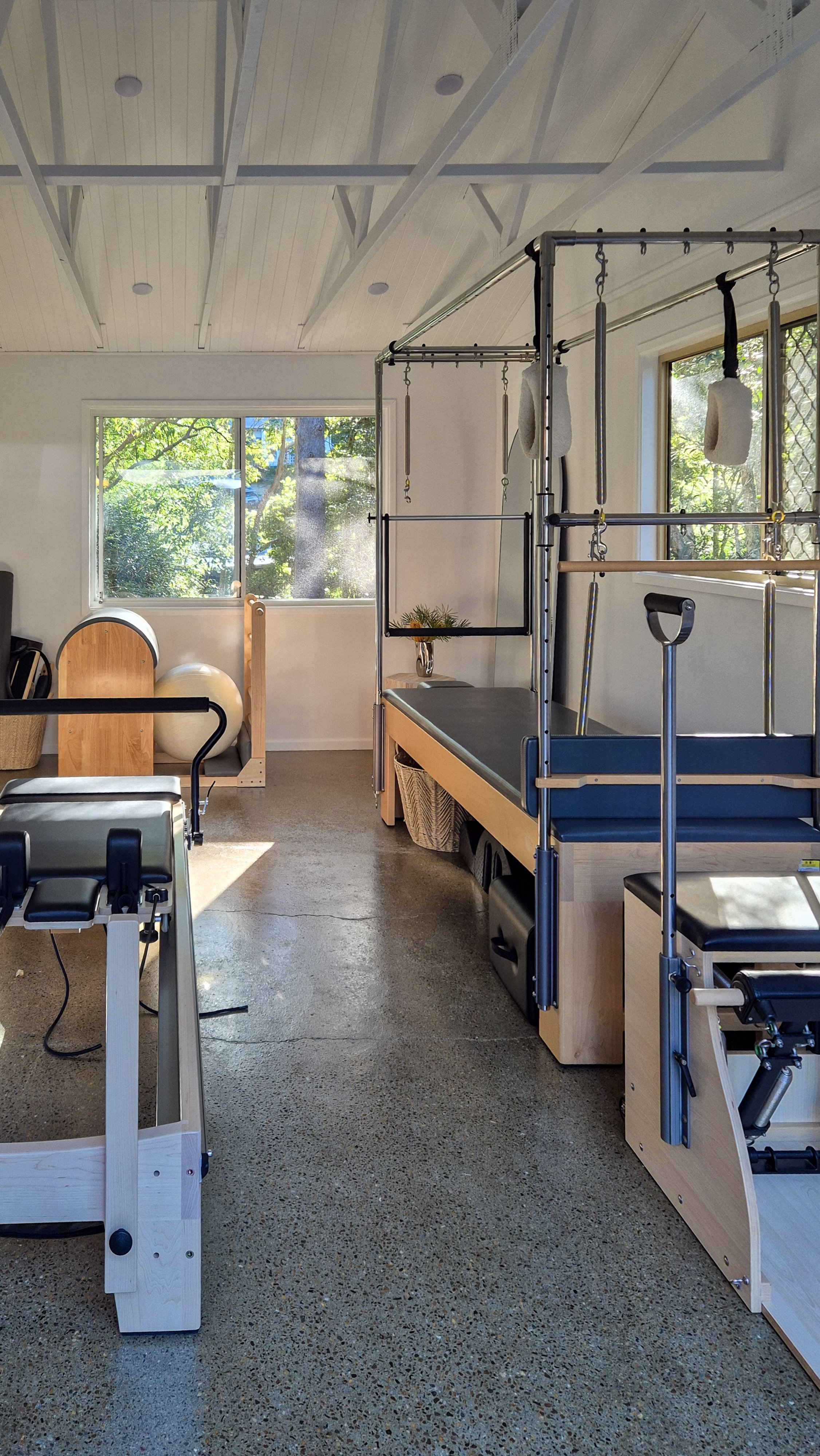 A therapy room with large windows showing trees outside, featuring A-frame wooden structures, fitness equipment, and a small elevated platform with rings and bars for physical therapy exercises.