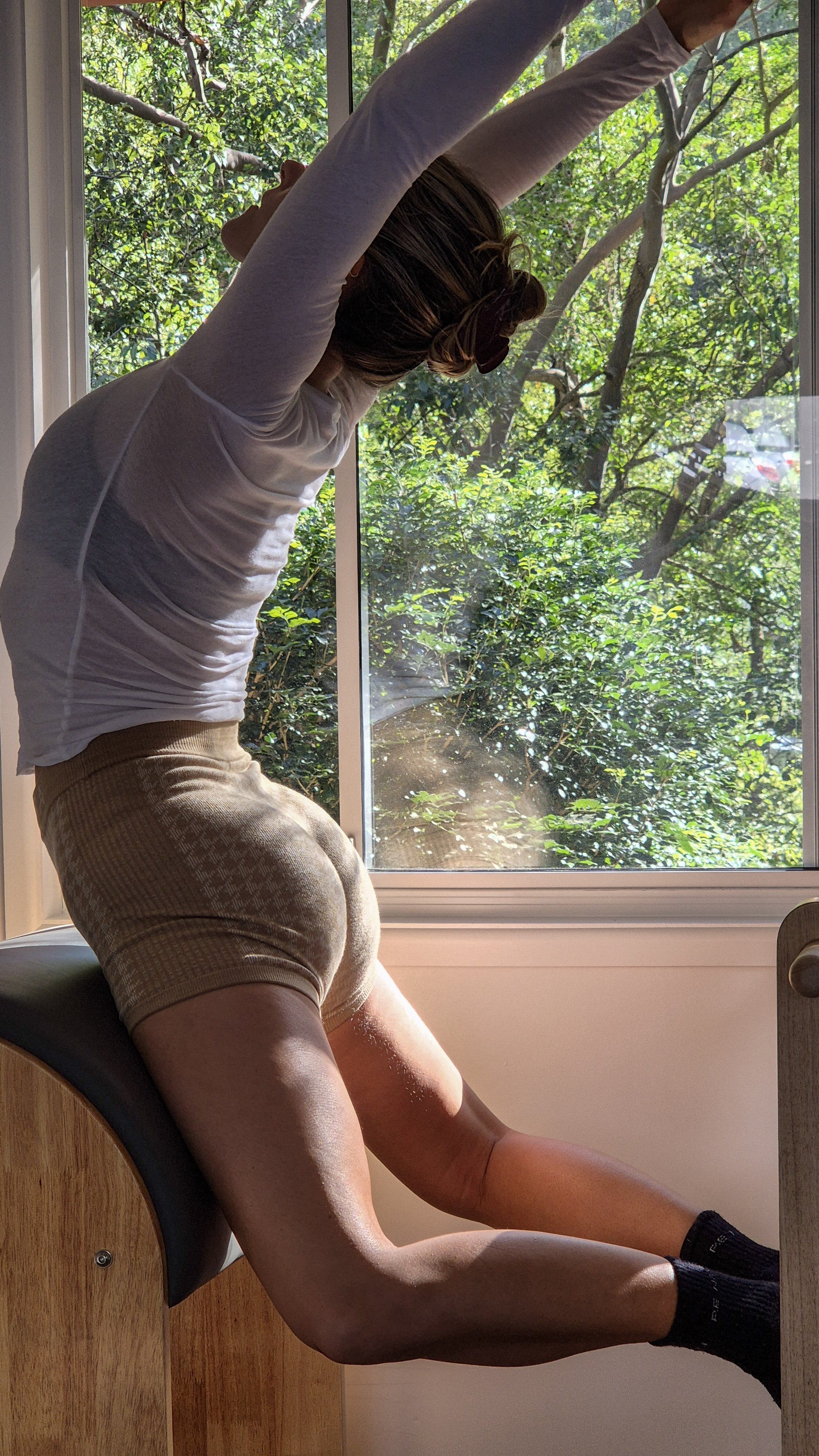 A person sitting on a mat near a window, stretching with arms raised overhead. They are wearing a long-sleeve shirt, beige shorts, and sunglasses, with greenery visible outside the window.