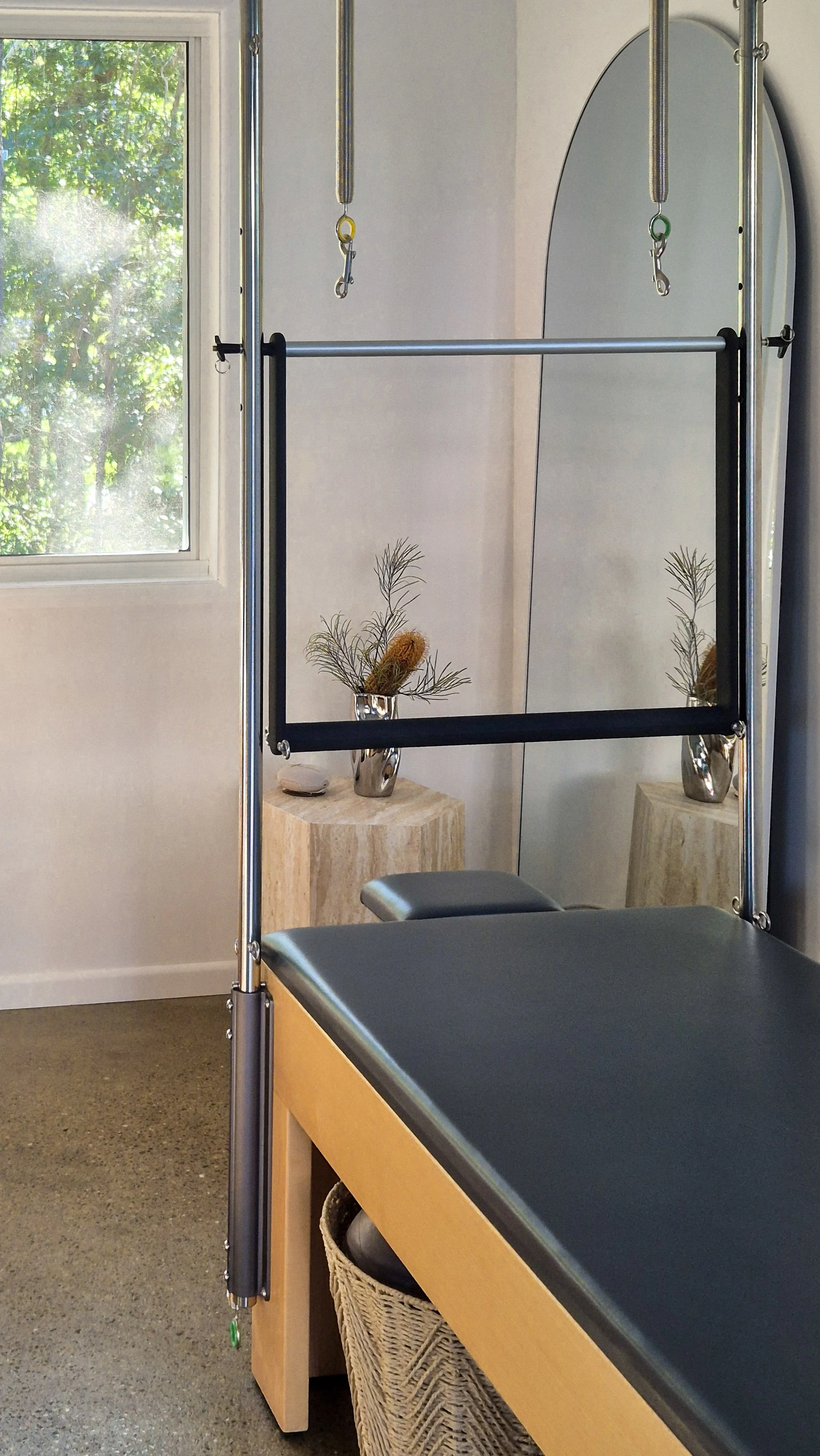 A physical therapy or exercise room with a doorway, a mirror, and a therapy table with black padding. There are two small tables with decorative plants in vases near a window with green foliage outside. Resistance bands with hooks are hanging from a metal frame.