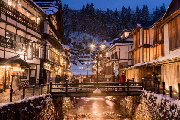 A Japanese onsen town with traditional 3-story wooden architecture along a stream with a foot bridge lit by the night light and street lamps while snowing in Ginzan Onsen, in Yamagata Japan.