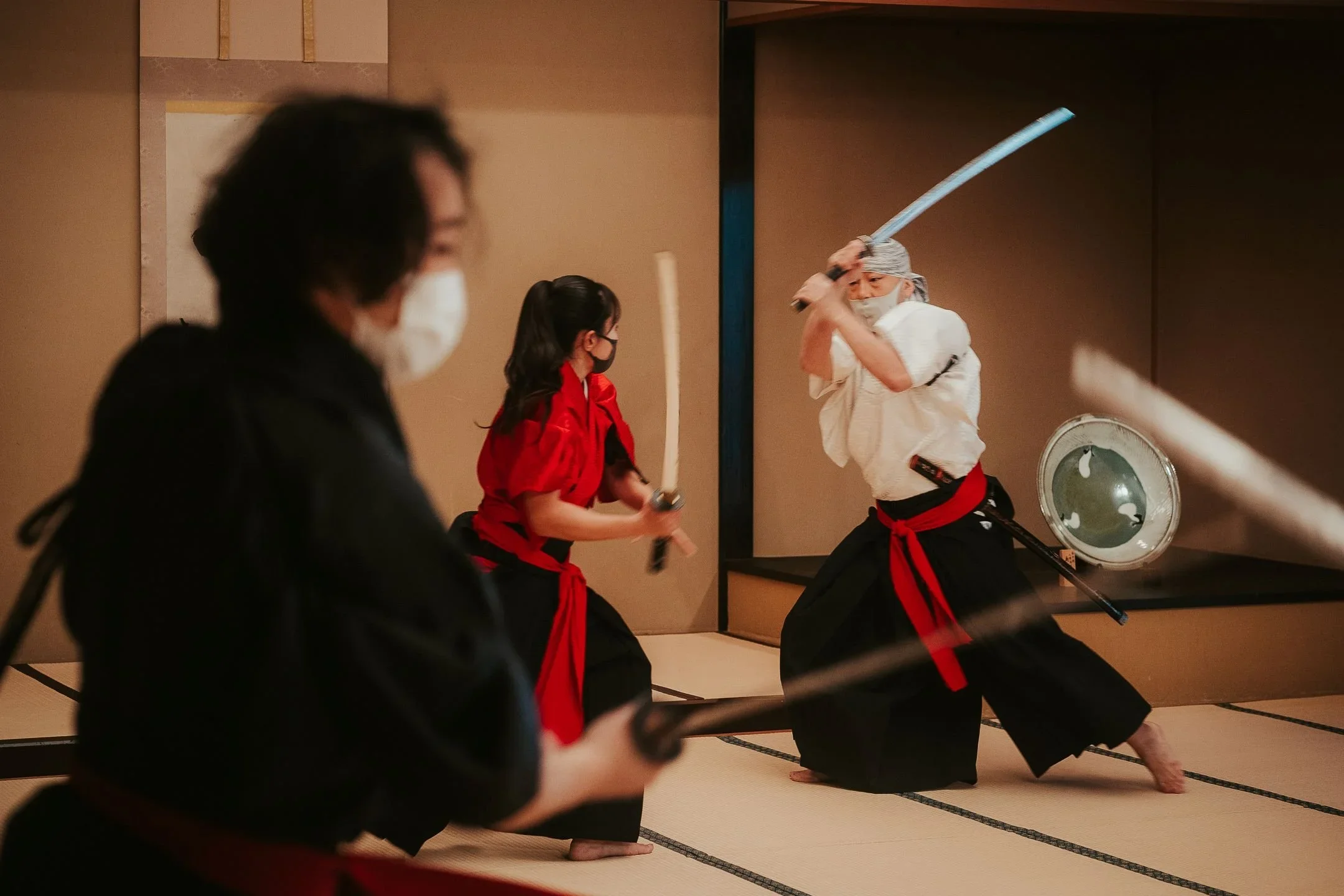 Man and woman wearing traditional Japanese kimono and hakama clothing fight with swords inside a tatami mat room.