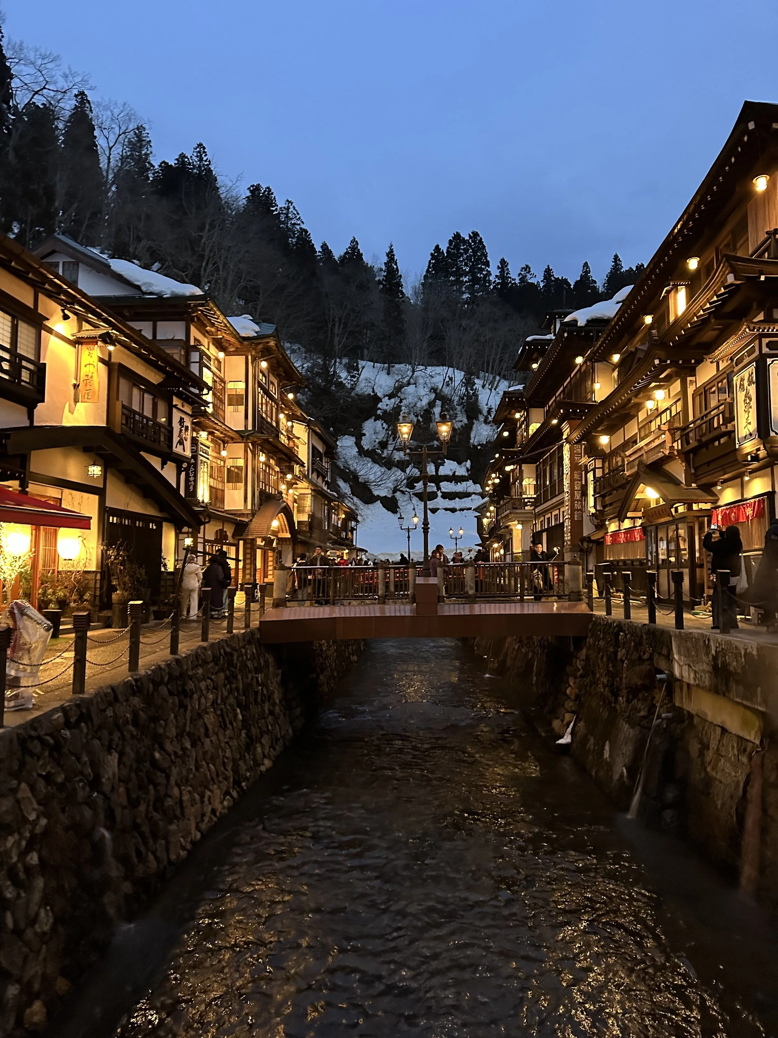 A scenic view of a Japanese village at dusk with traditional buildings illuminated by warm lights, a small river flowing underneath the bridge, snow on the rooftops and trees, and a mountain with trees in the background.
