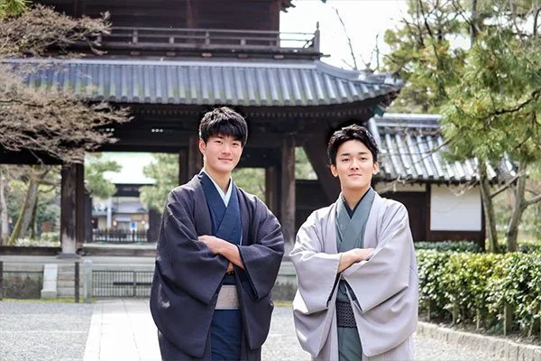 Two young men stand with their arms folded in front of a wooden temple entrance dressed in a blue and a grey and green kimonos in Japan.