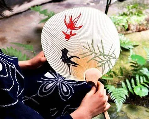 A fan painted with goldfish held by someone in a blue yukata in a lush landscape.