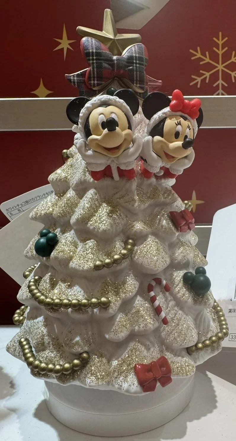 Mickey and Minne Mouse decorate a white christmas tree with a gold gardland and red bows and green mickey shaped bulbs figure.