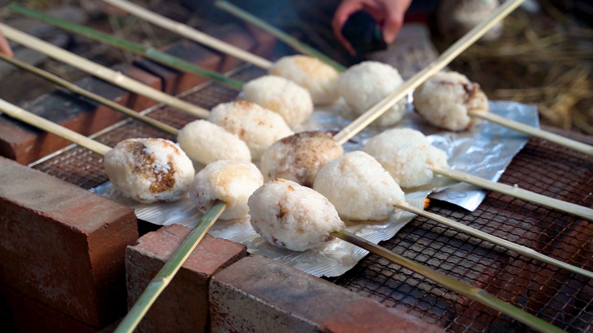 12 gohei mochi balls on sticks are roasting over a fire pit.