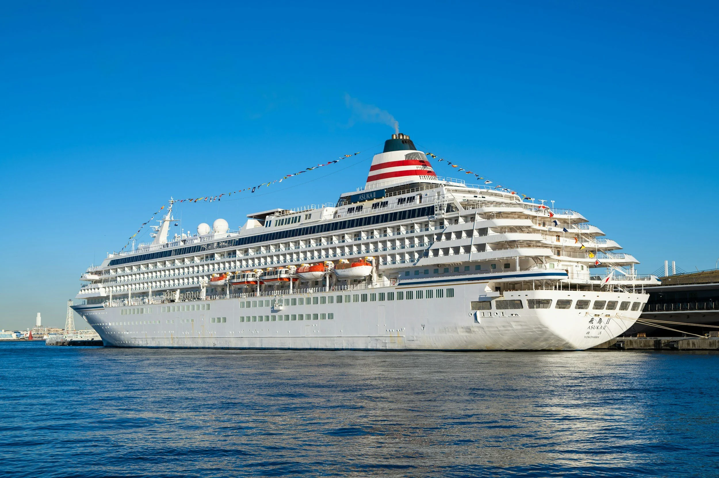 A large white cruise ship docked at a port under a clear blue sky, with small flags hanging from the top of the ship.