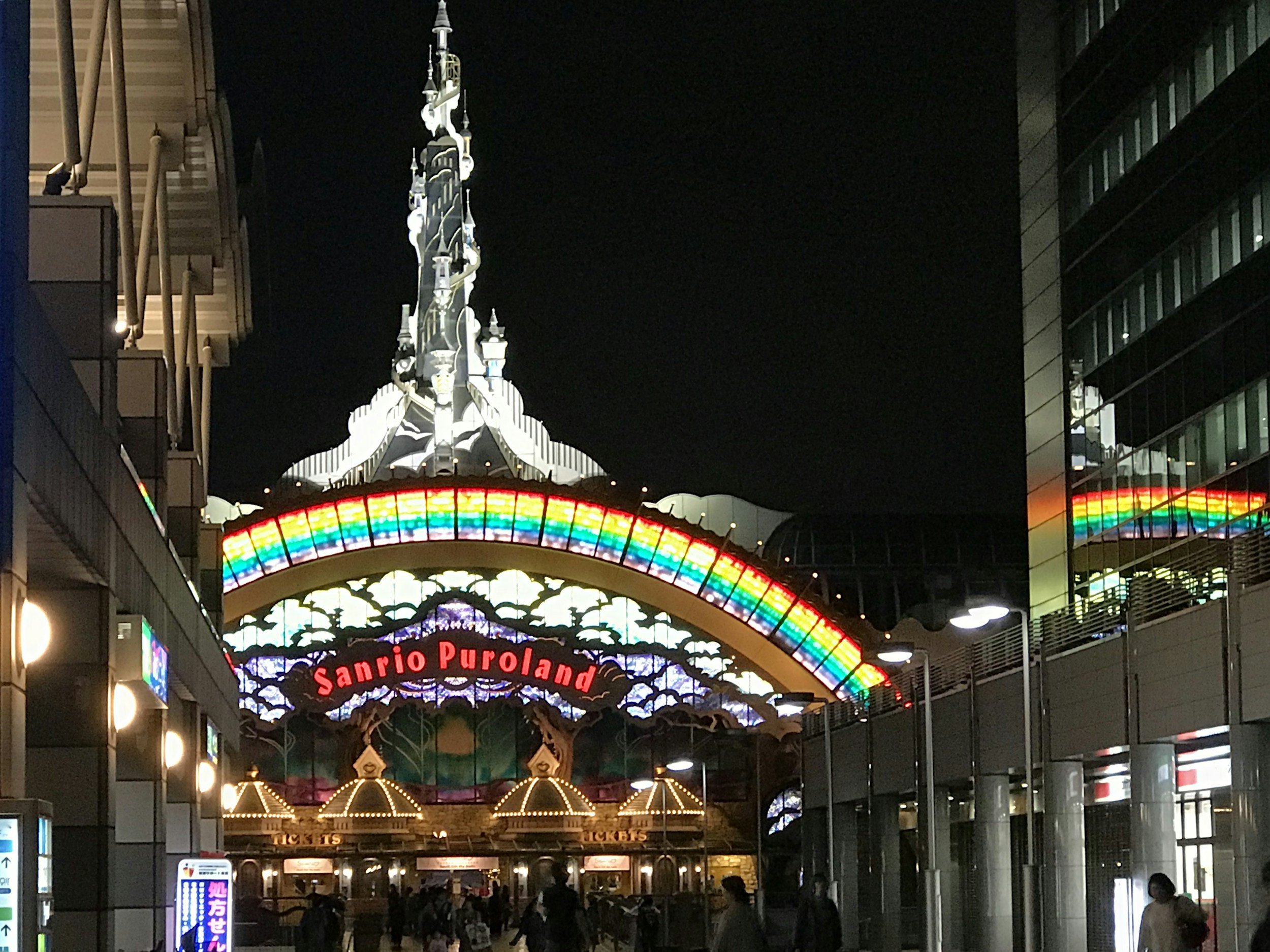 SanrioPuroland entrance way at night with a rainbow color archway and a castle like spire.
