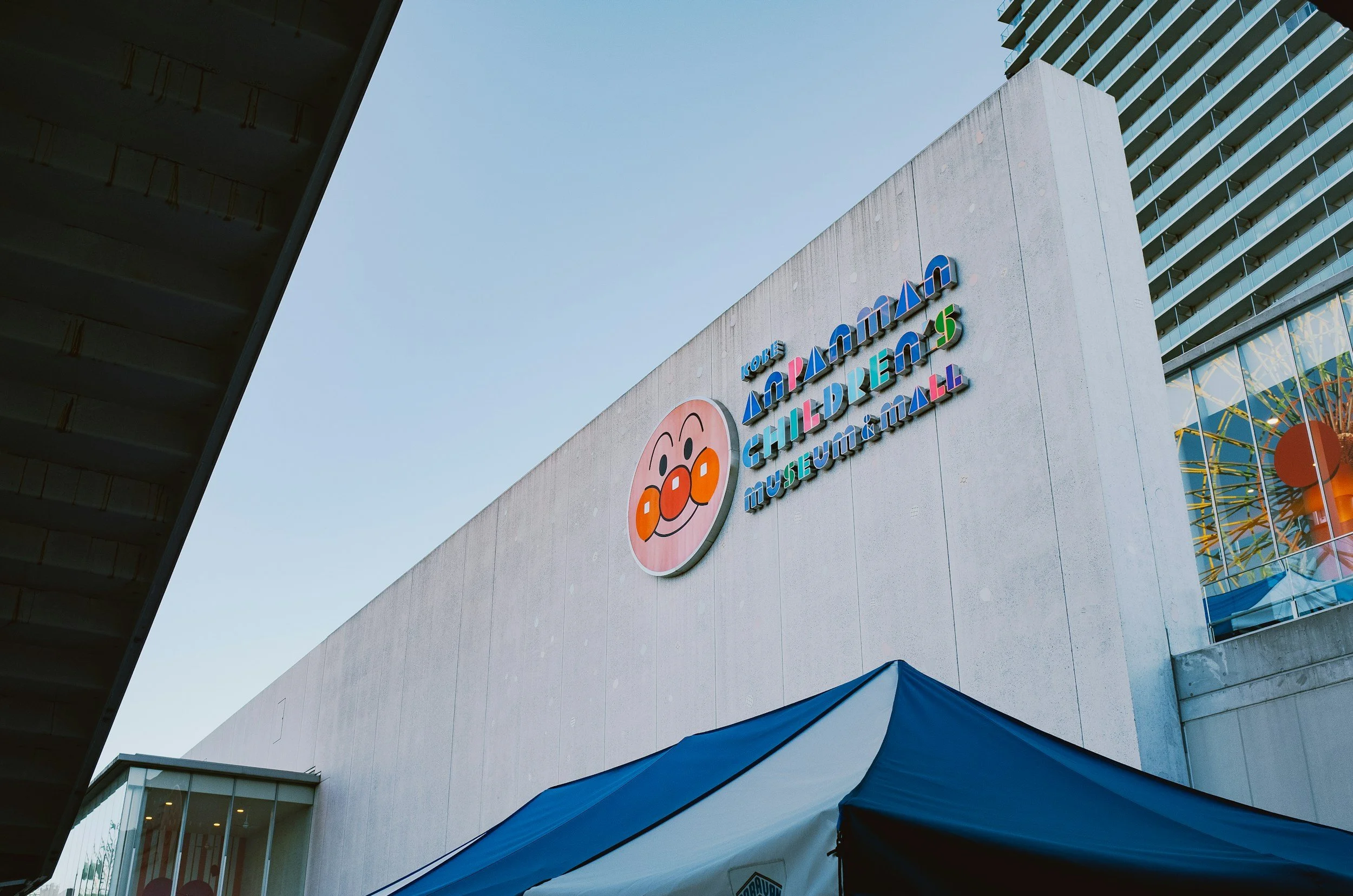 Colorful and geometric signage on the top of the gray concret building of the Anpanman museum with the face of Anpanman mounted to the wall taken from below the sign.
