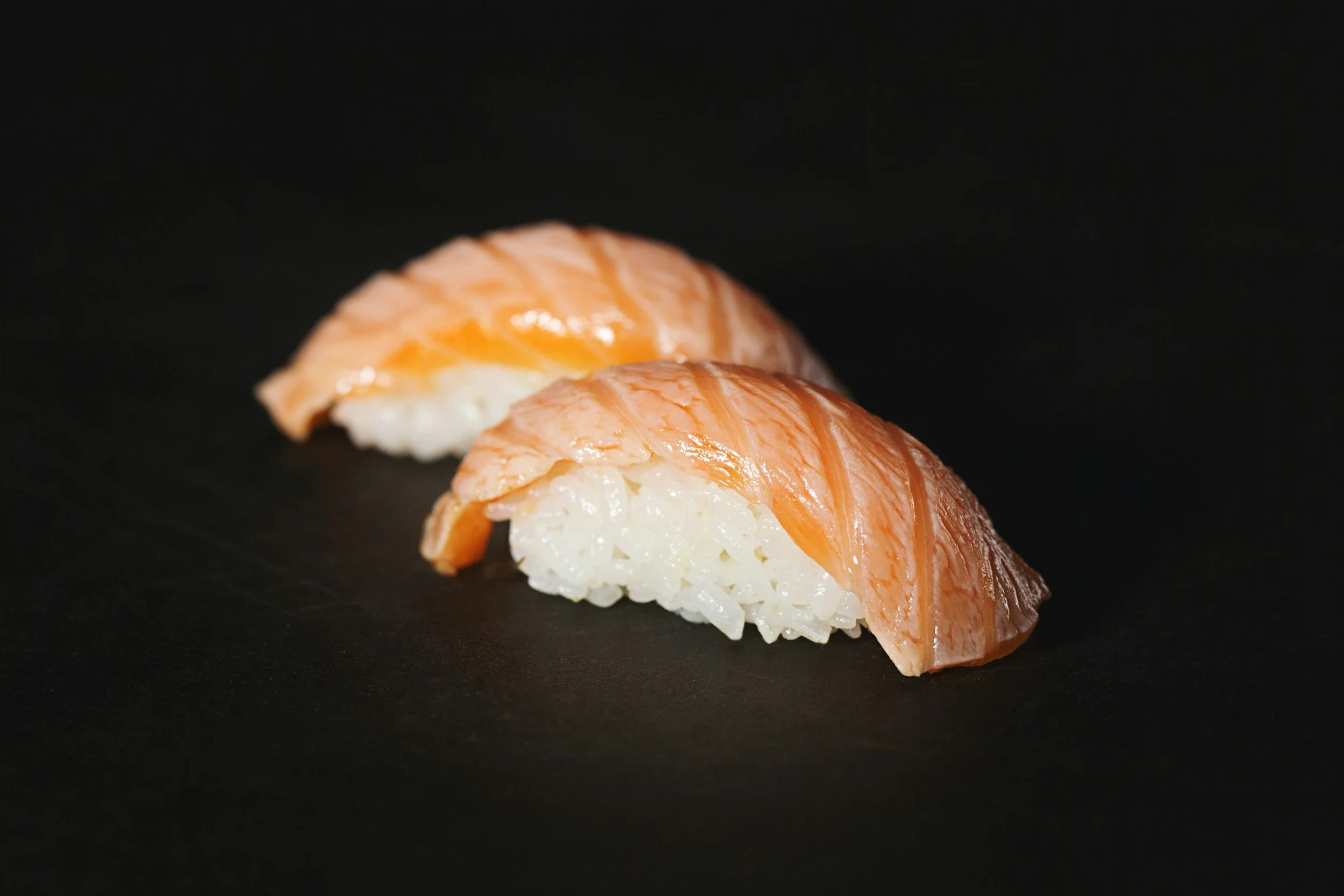2 pieces of salmon drape over a ball of vinegar rice to form a nigiri sushi roll resting on a black plate.