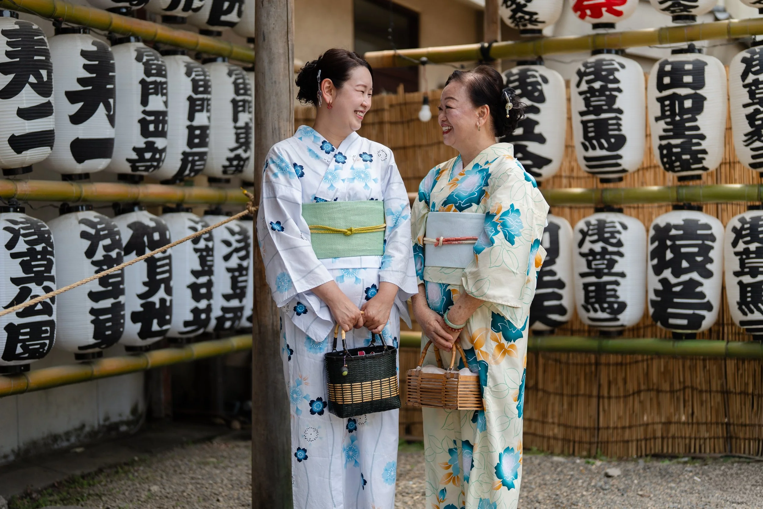 An asian mother and a daughter partake in a light colored yukata dressing experience stand in a garden look at each other surrounded by Japanese lanterns.