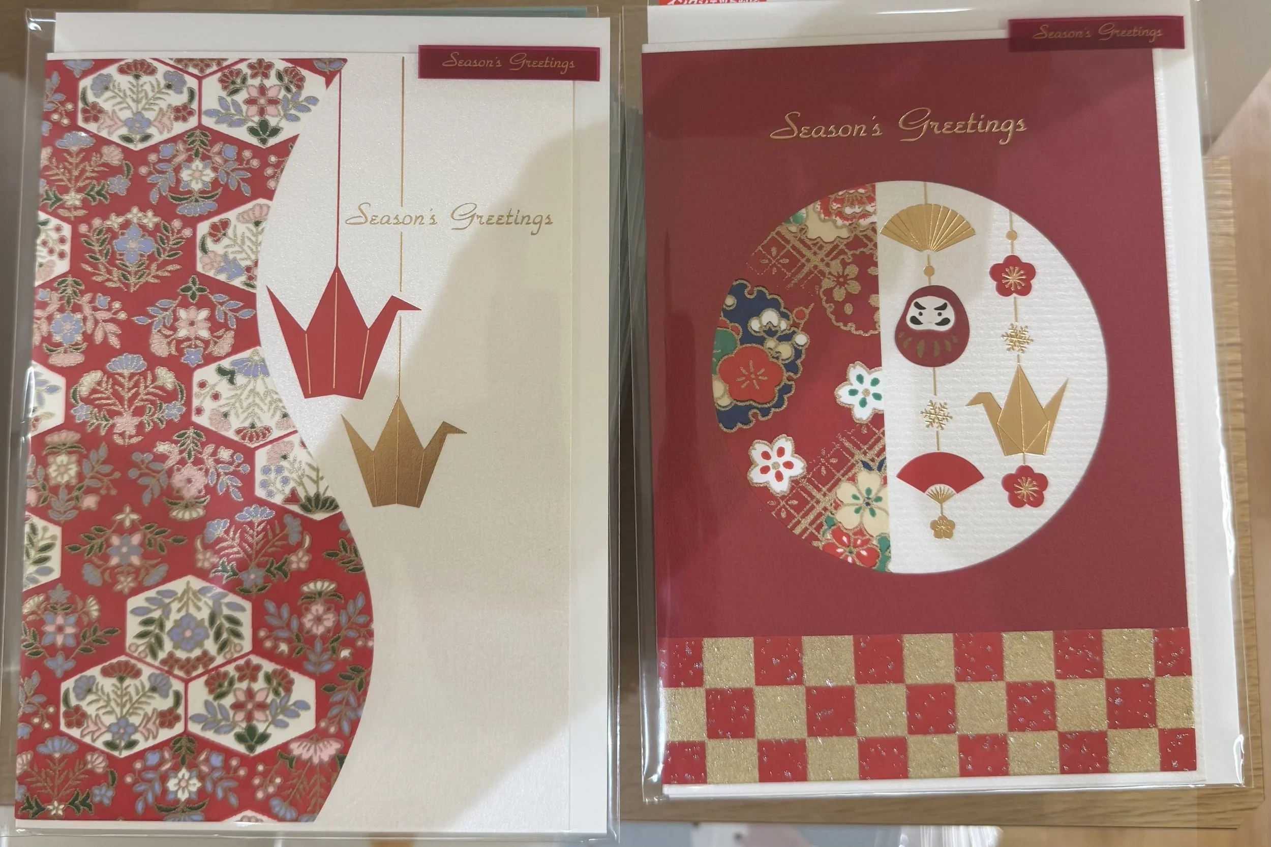 Two red Christmas and seasonal greeting cards with Japanese moifs like a fan, crane, and daruma doll sit next to each other.